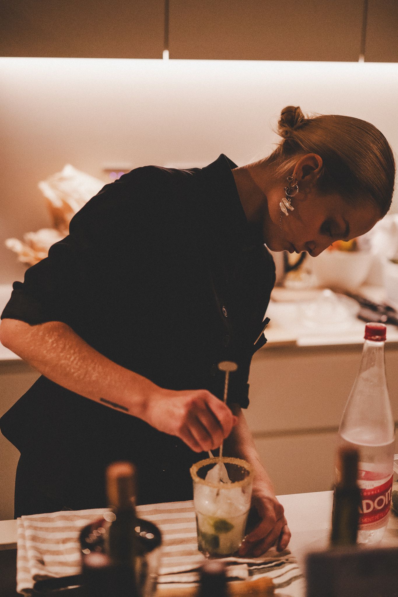Female bartender in black outfit carefully preparing a cocktail drink at a warmly lit bar counter.