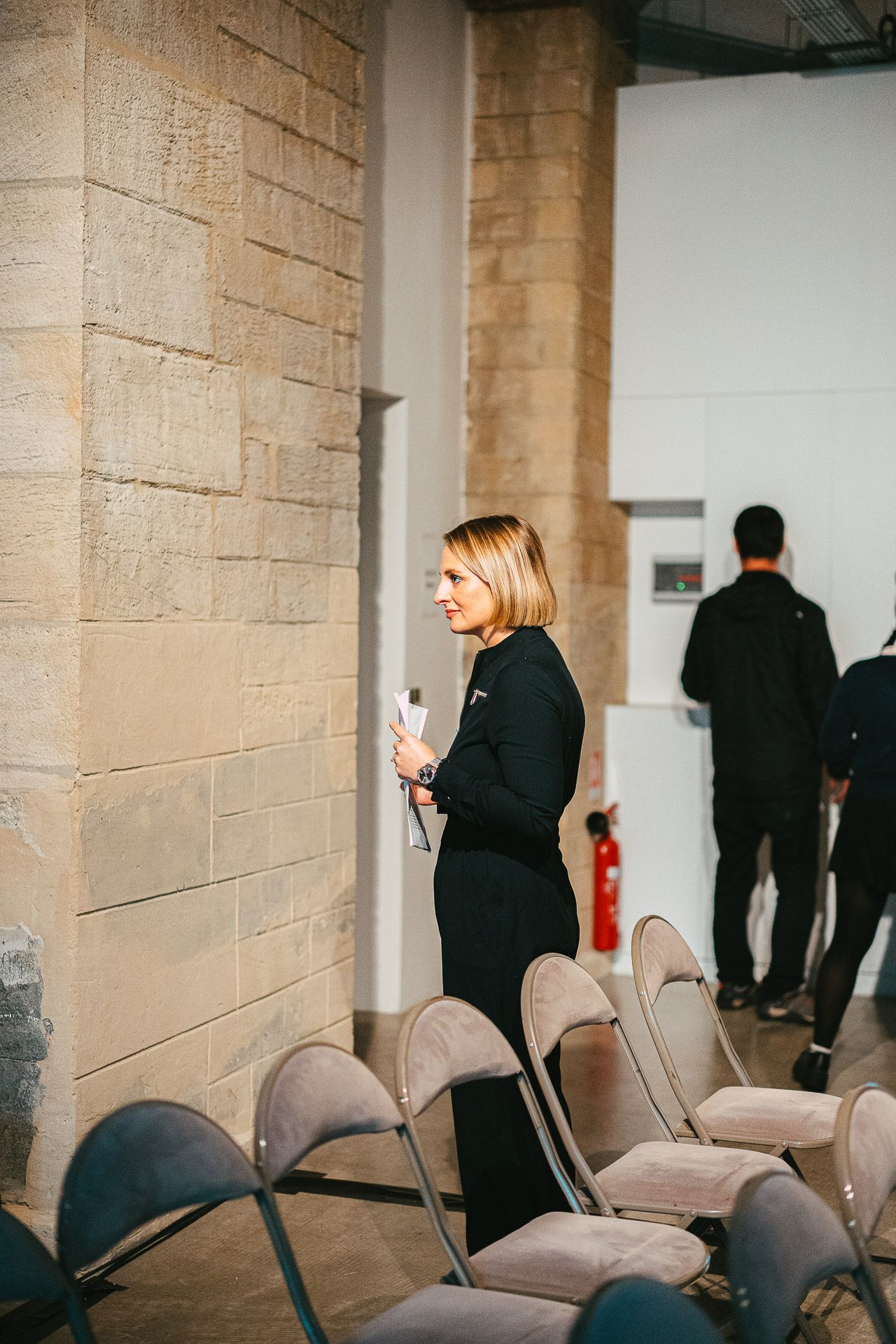 Blonde woman in black dress holding microphone standing among empty chairs in stone venue.