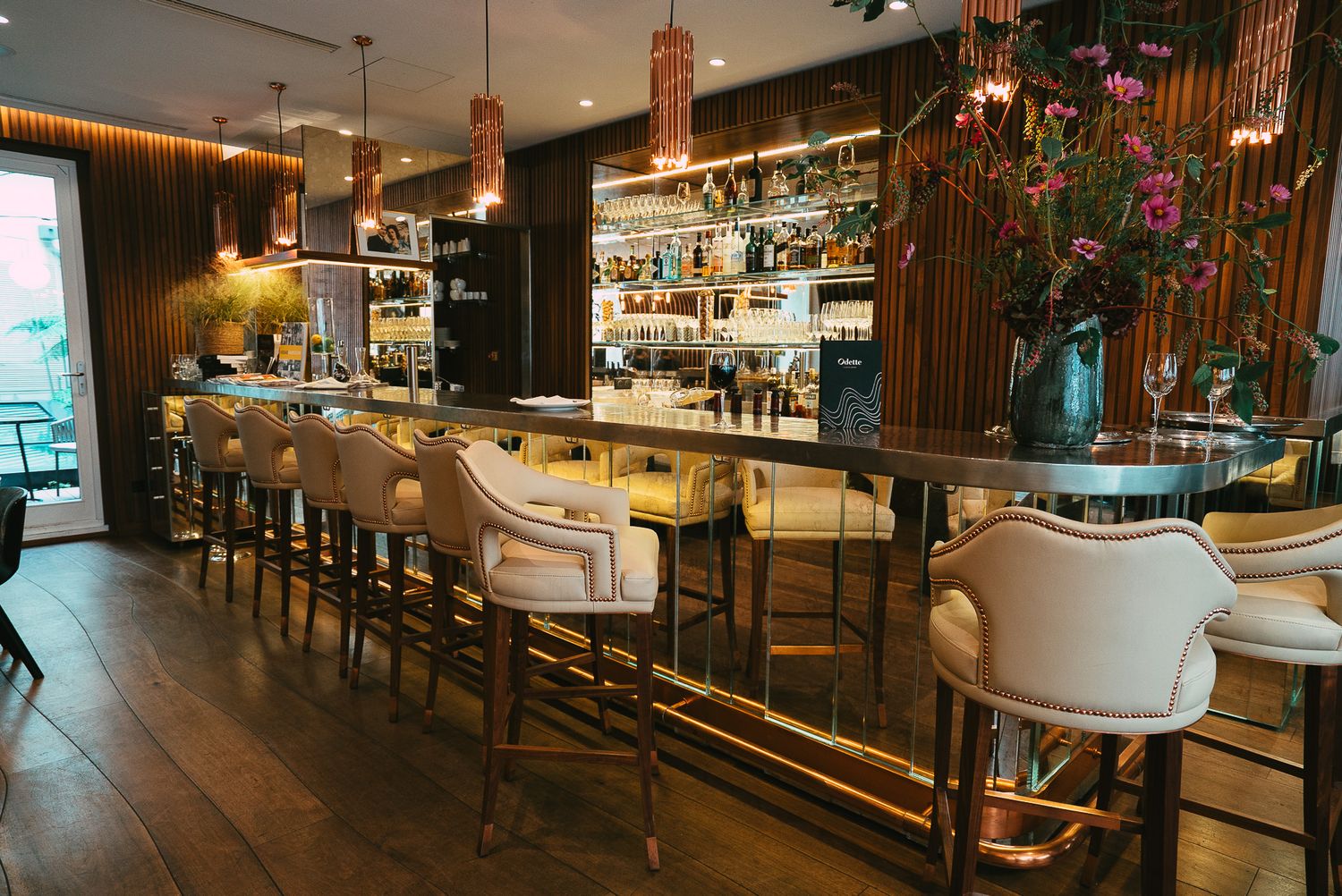 Elegant restaurant bar with cream upholstered stools, warm wood paneling, pendant lights, and fully stocked back bar.