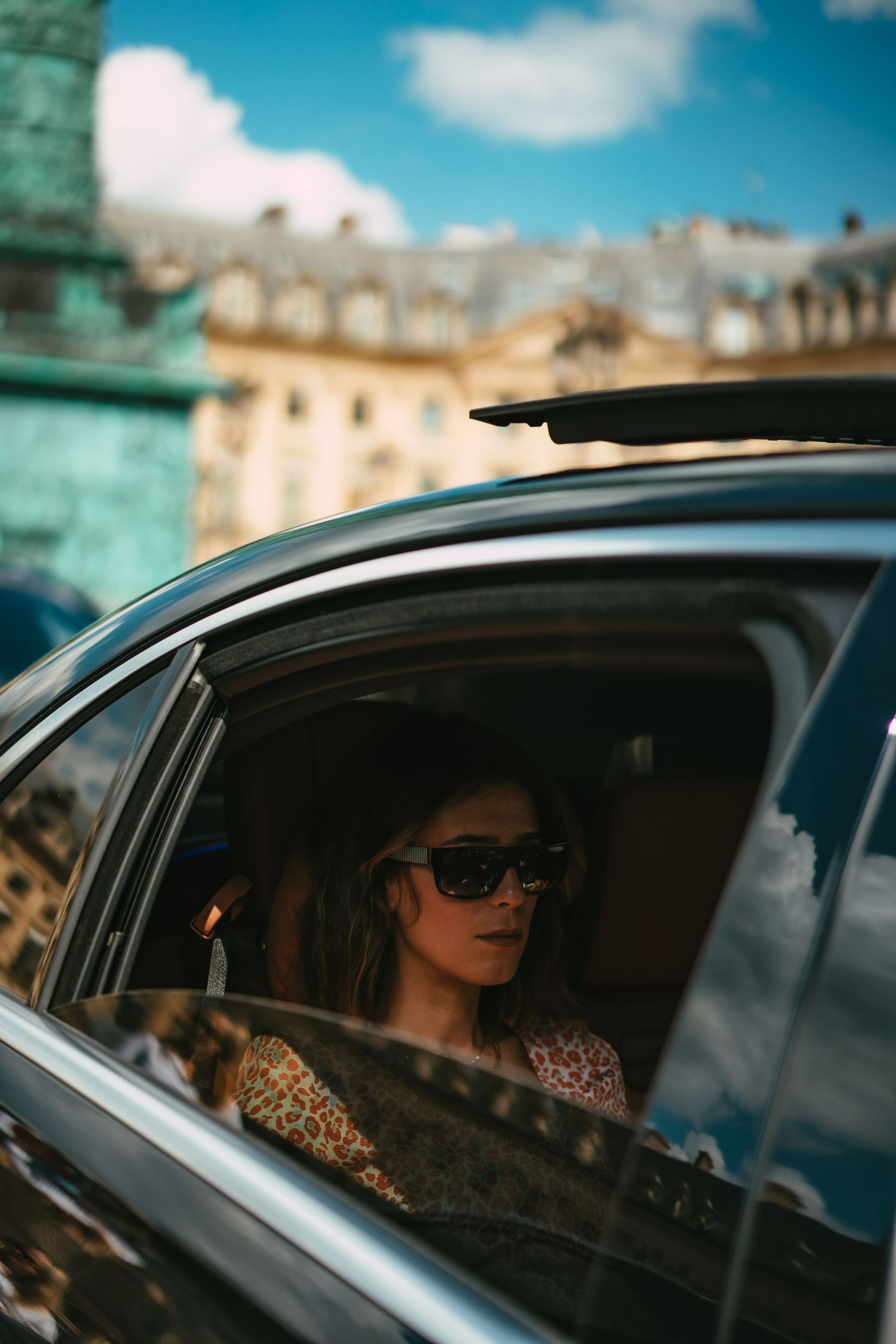 Woman in sunglasses and floral top seated in luxury car backseat in Paris.