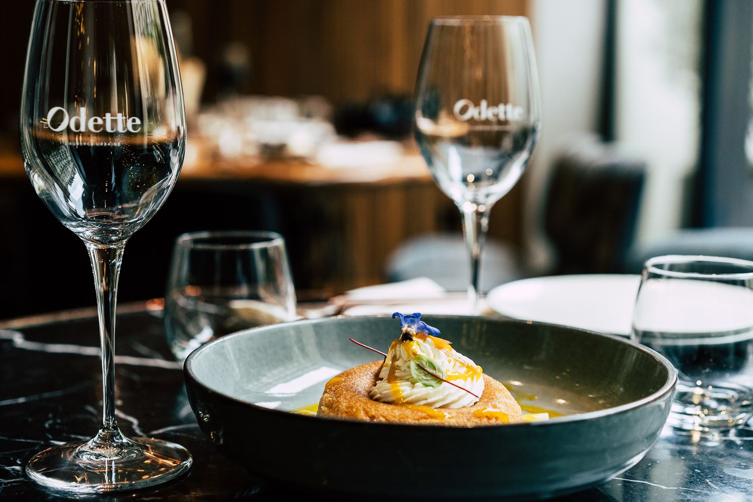 Elegant fine dining dish served in a gray bowl at Odette restaurant with branded wine glasses on marble table.