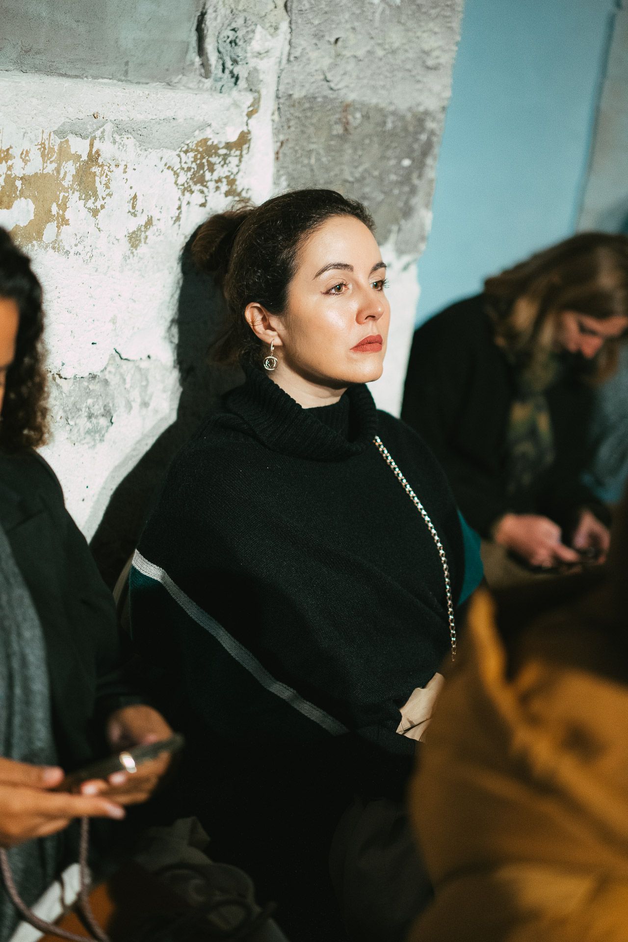 Woman in black turtleneck sweater with red lips standing against a rustic wall at event.