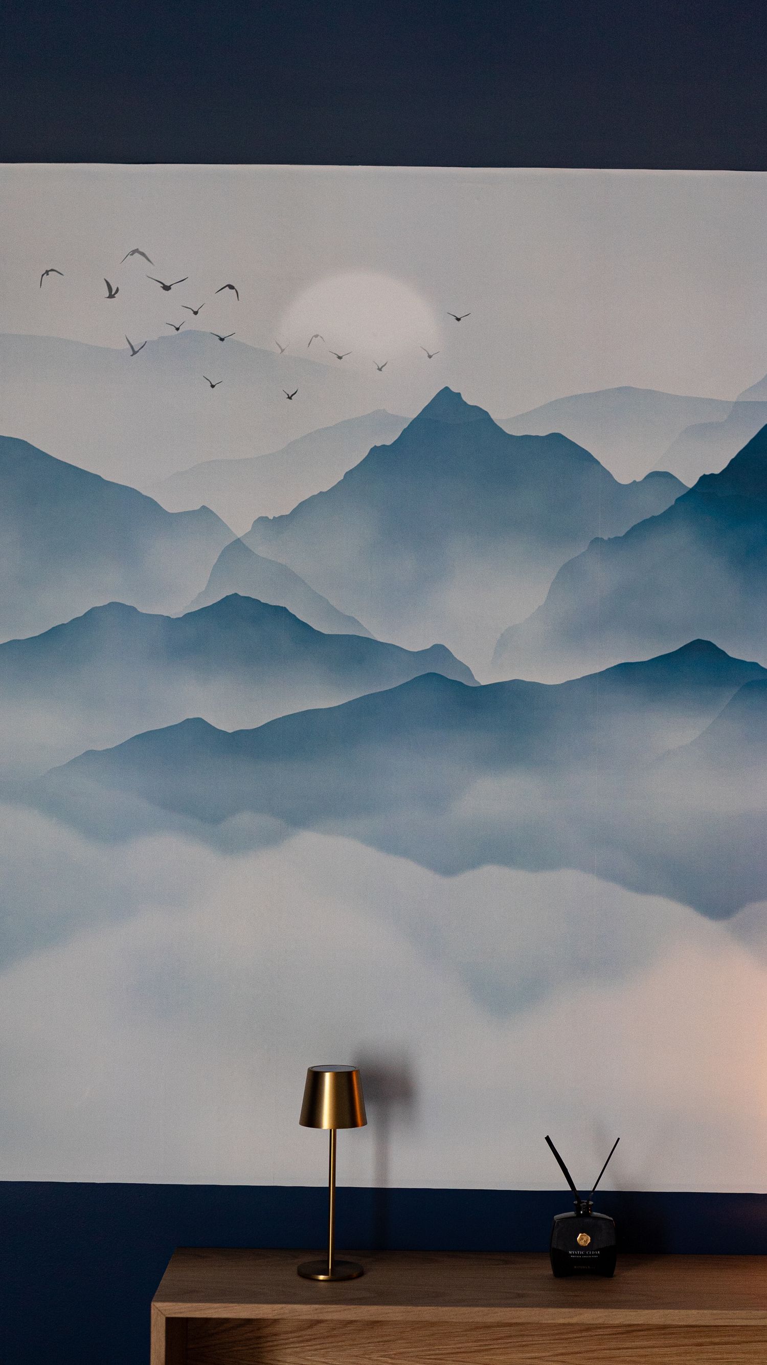 Serene blue mountain landscape mural wallpaper with misty peaks, birds, and small brass table lamp on wooden shelf.