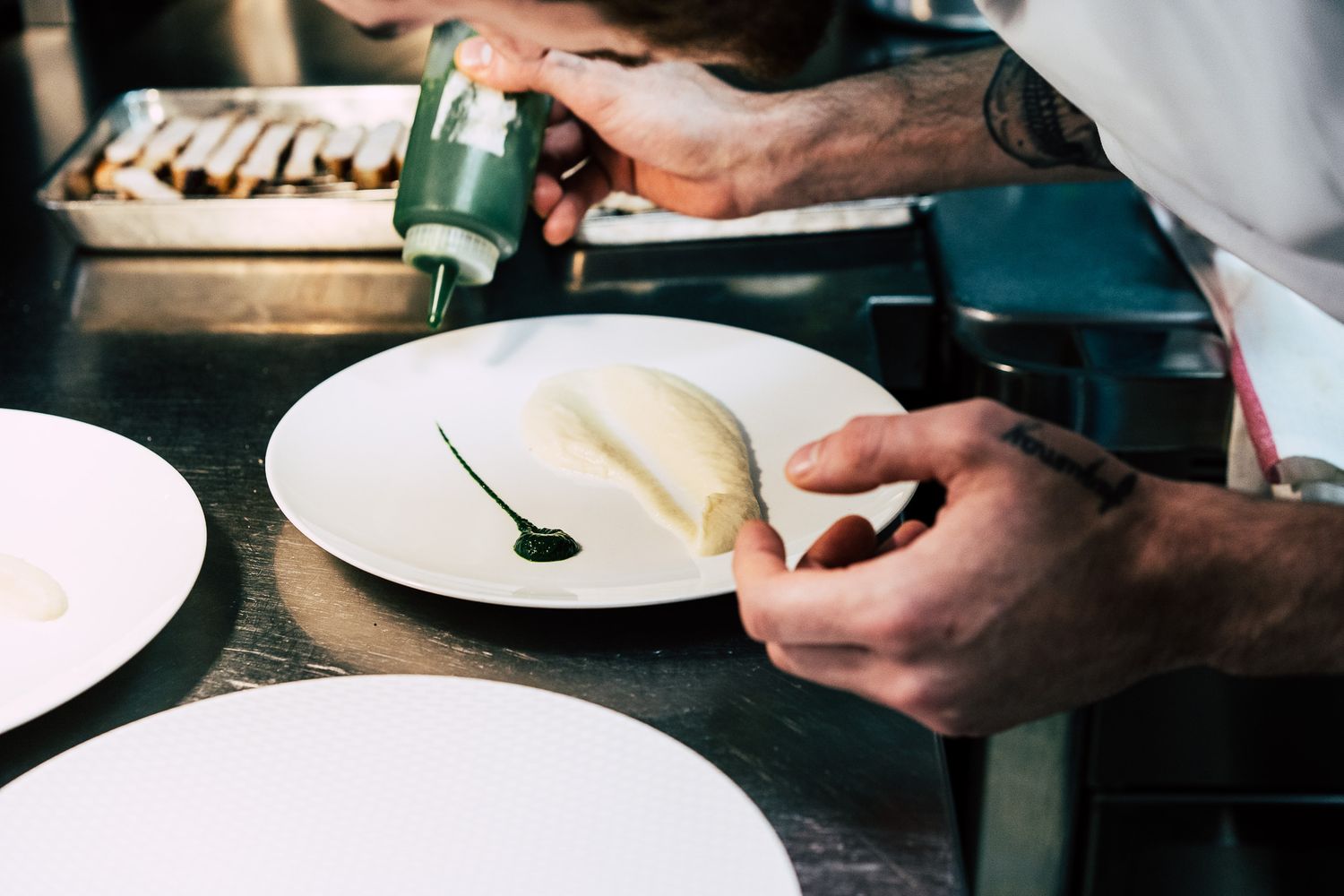 Chef plating an elegant dish with green sauce on a white plate in a restaurant kitchen.