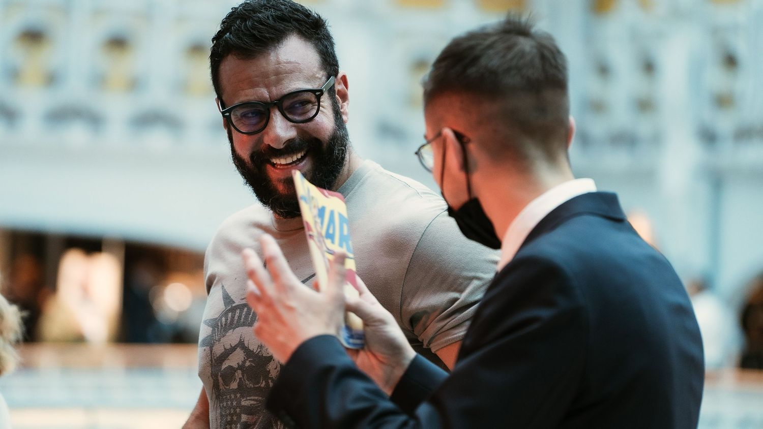 Two men in conversation at an event, one with tattoos and glasses, the other wearing a face mask.