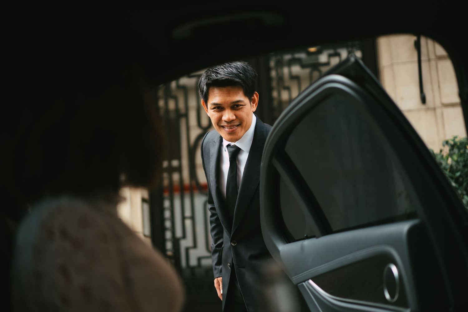 Smiling young Asian man in suit holding open a car door, viewed from inside the vehicle.