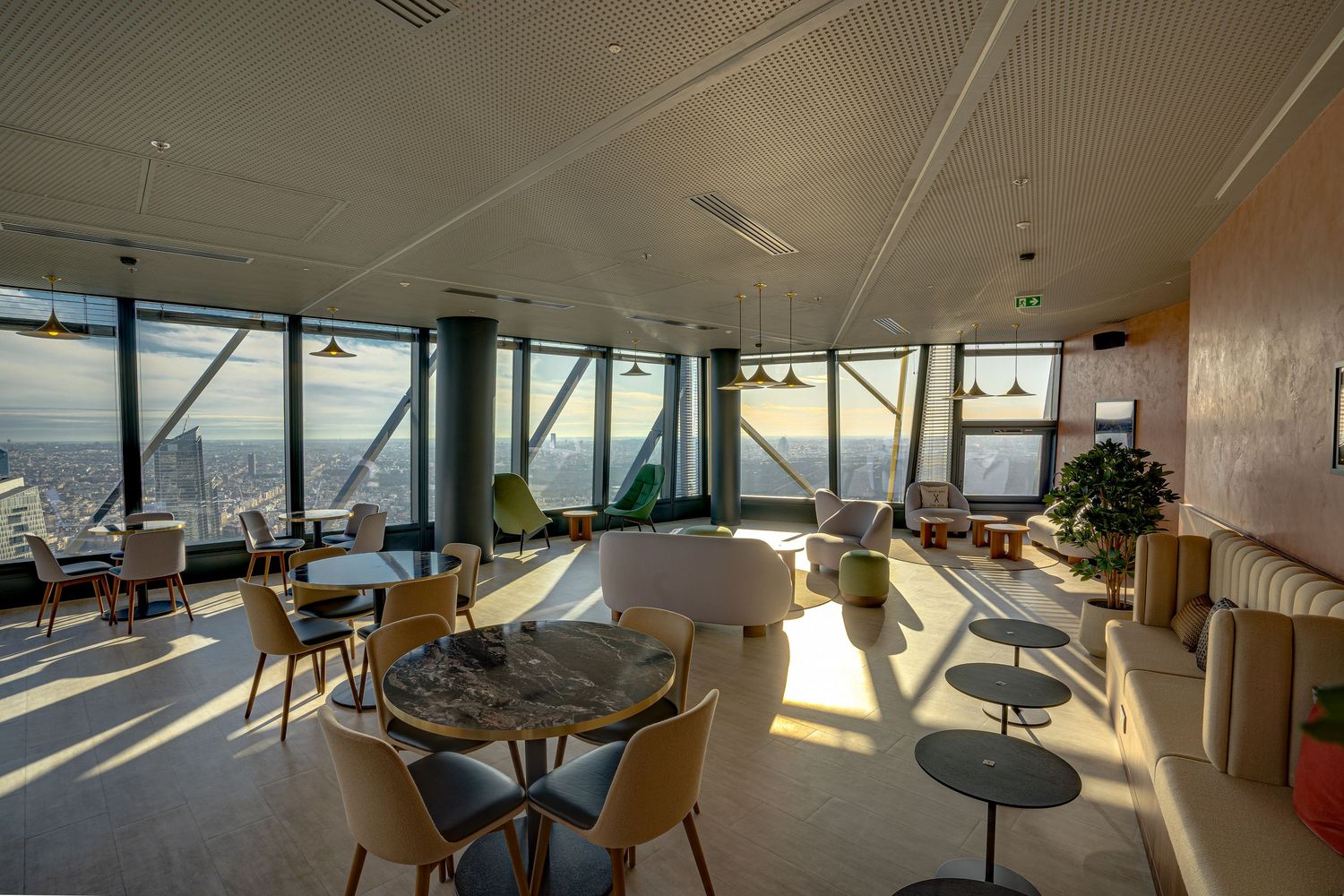Sunlit high-rise lounge with panoramic city views, round tables, colorful chairs, and modern decor.