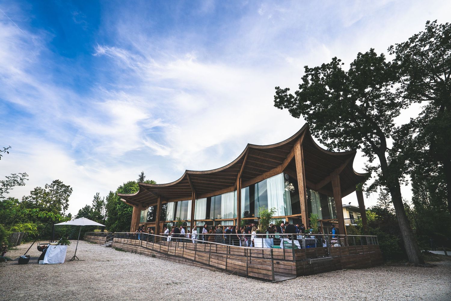 Modern curved glass pavilion restaurant surrounded by trees under a blue sky with guests dining outside.
