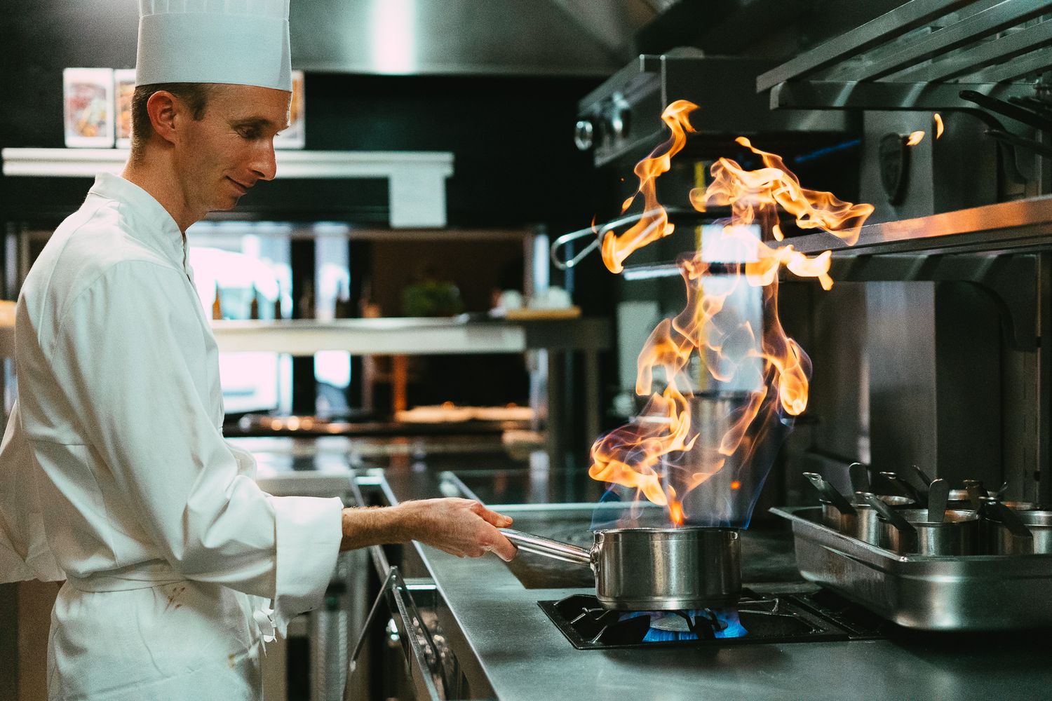 Smiling chef in white uniform flambeing food with high flames in professional kitchen.
