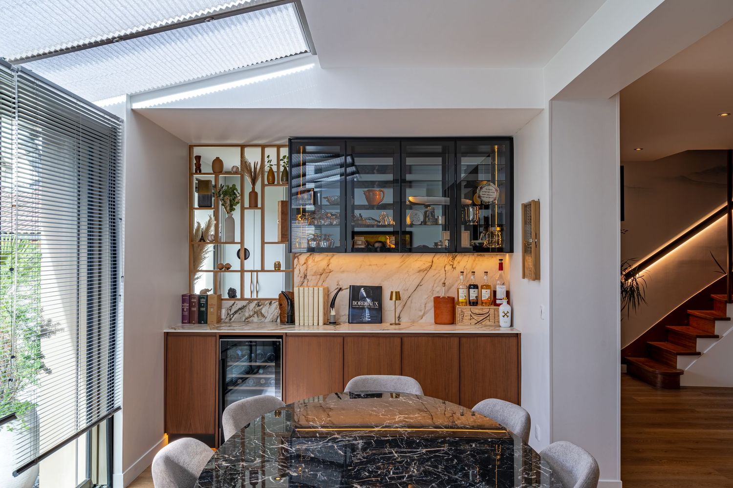 Stylish dining area featuring dark marble table, walnut bar cabinet, glass shelving, and staircase with warm lighting.