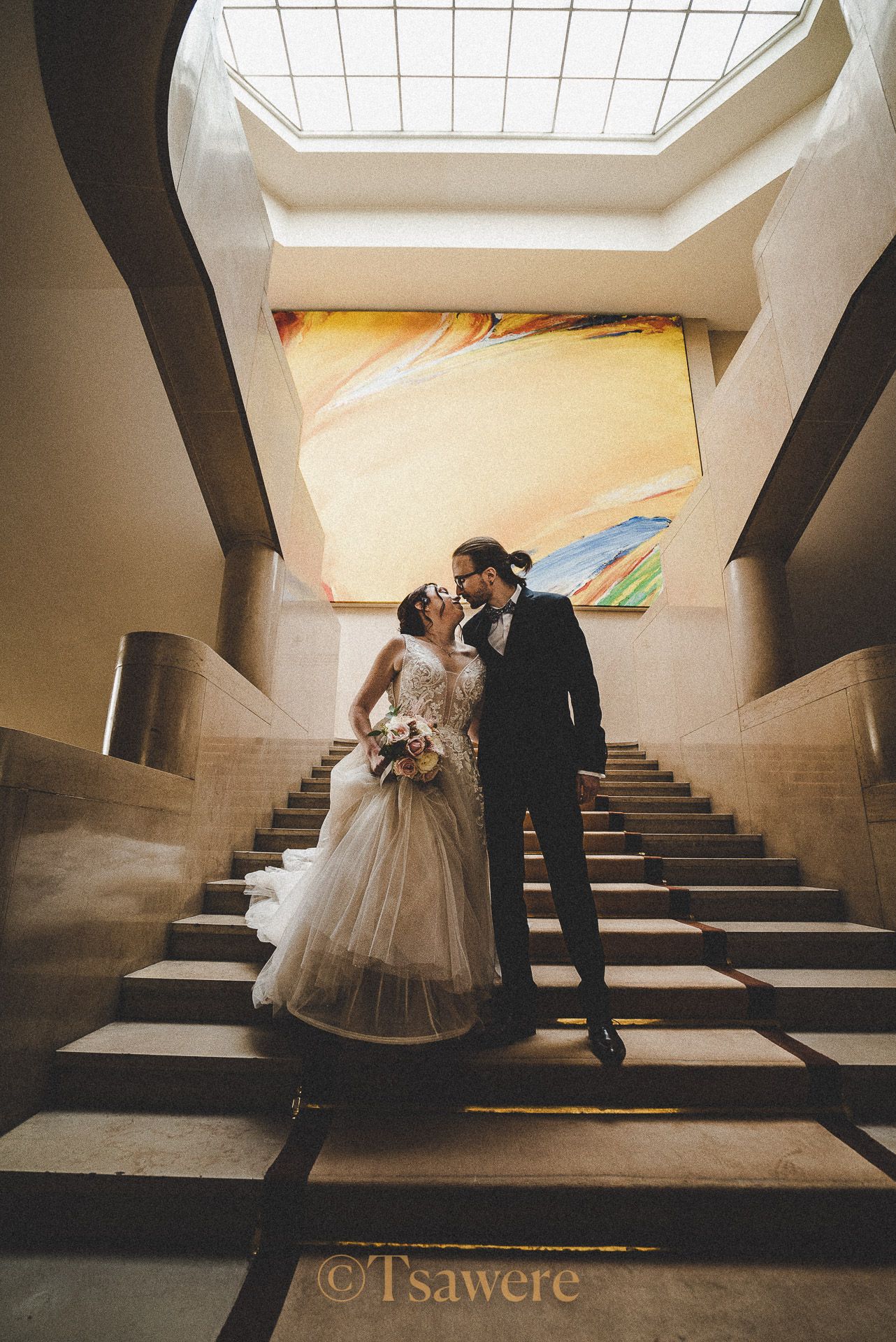 Bride and groom sharing a kiss on grand staircase beneath skylight in elegant building.