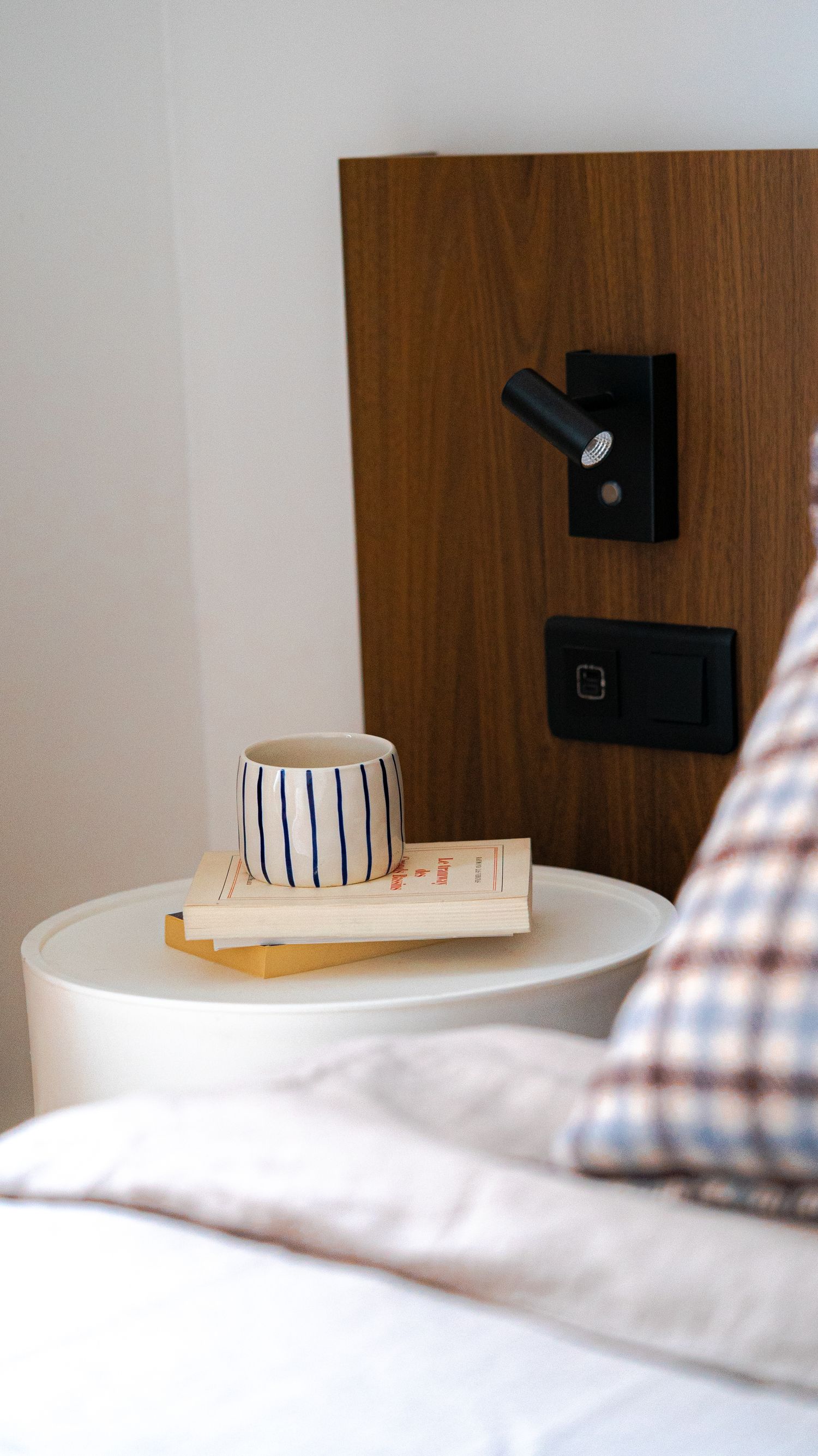 Minimalist bedside table with striped ceramic mug, books, and black wall-mounted reading light on wooden headboard.