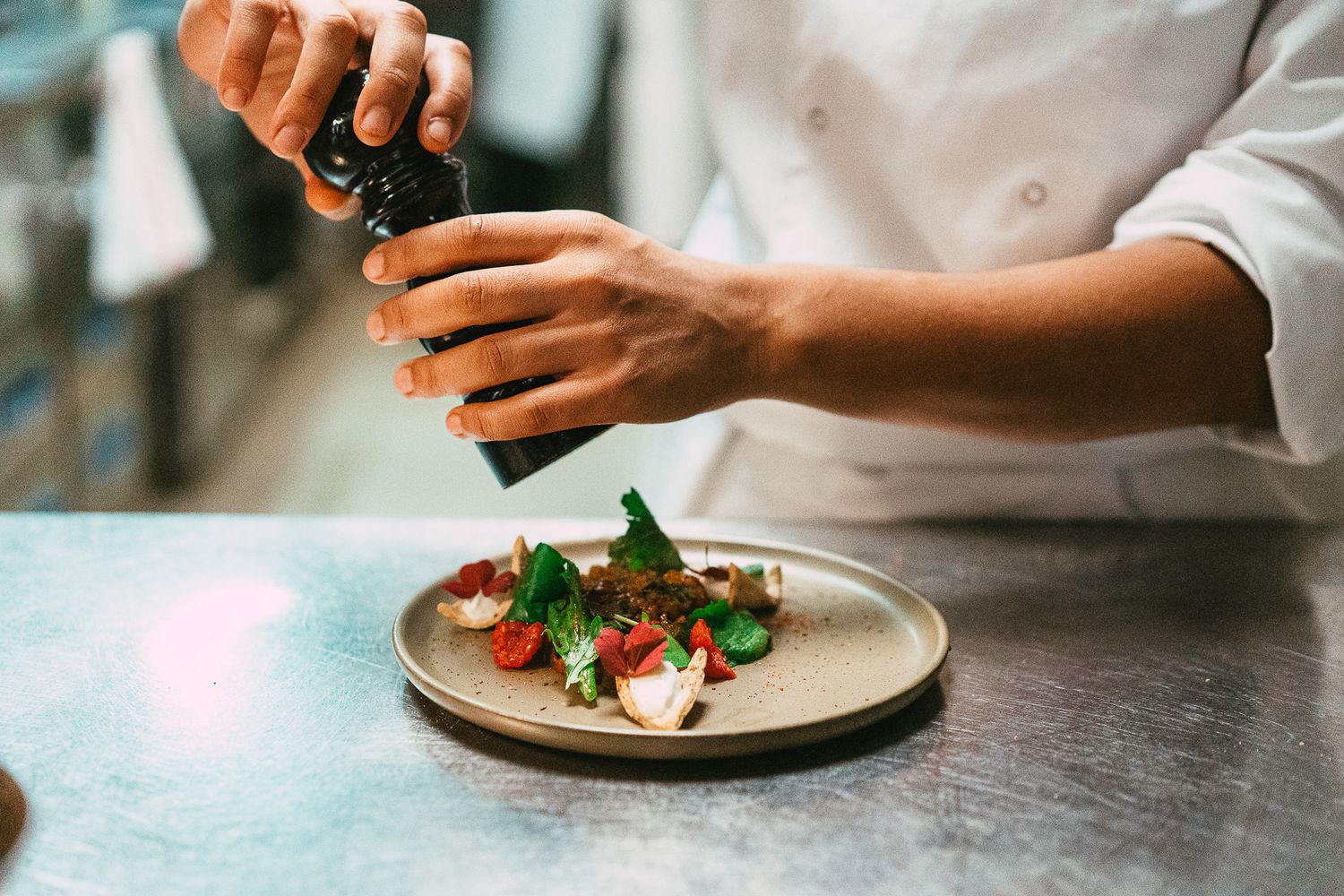 Chef grinding black pepper over a gourmet dish with fresh vegetables on a ceramic plate.