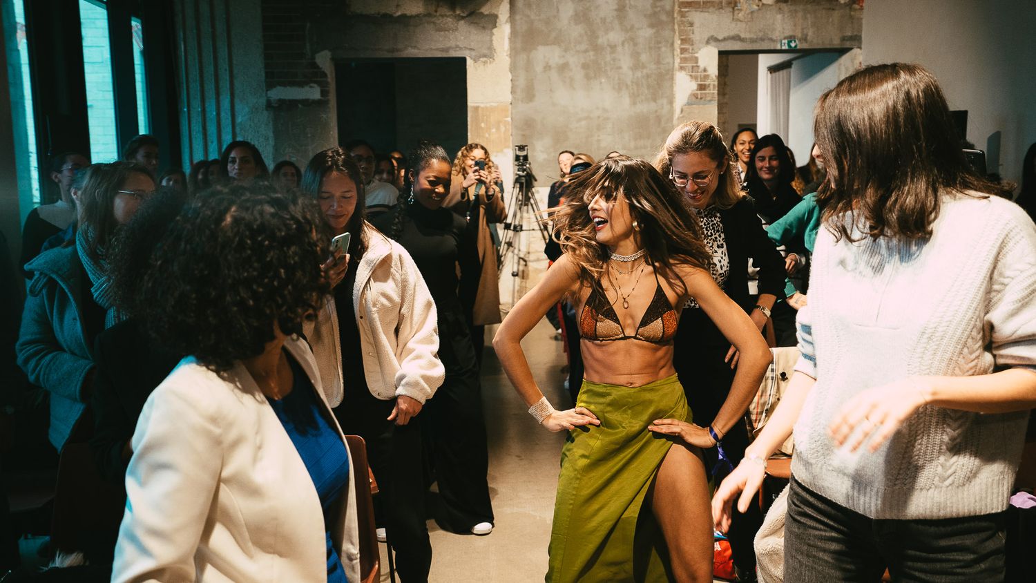 Woman in a bikini top and green skirt dancing at a crowded indoor fashion event.