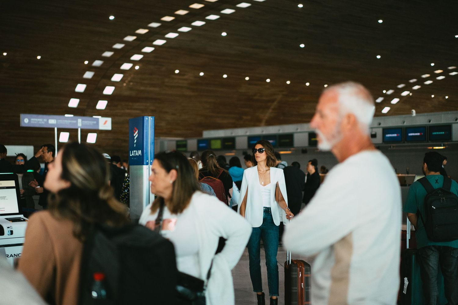 Woman in white blazer standing confidently in busy airport check-in hall surrounded by travelers.