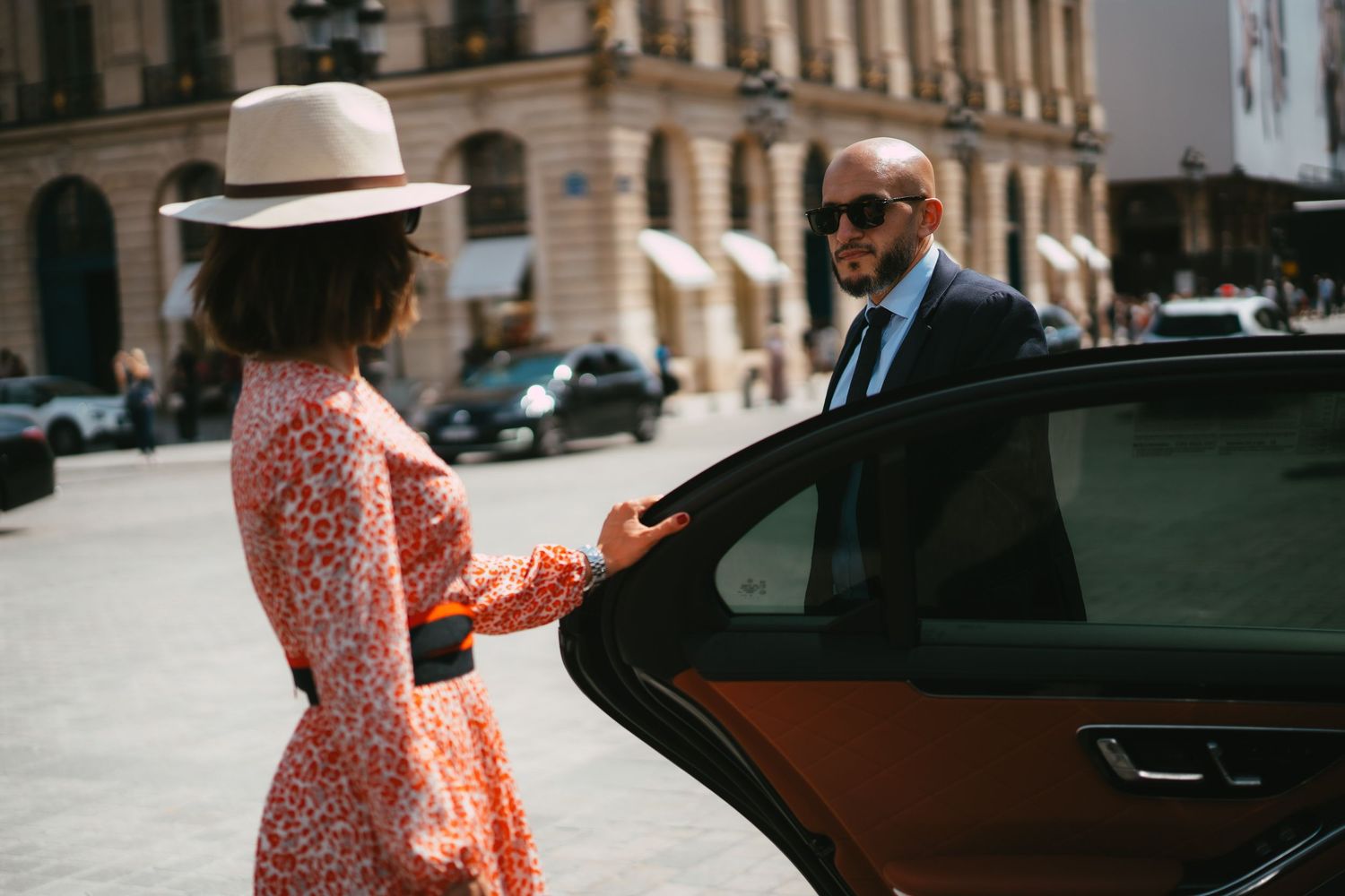 Chauffeur opening luxury car door for stylish woman in floral dress and hat on sunny street.