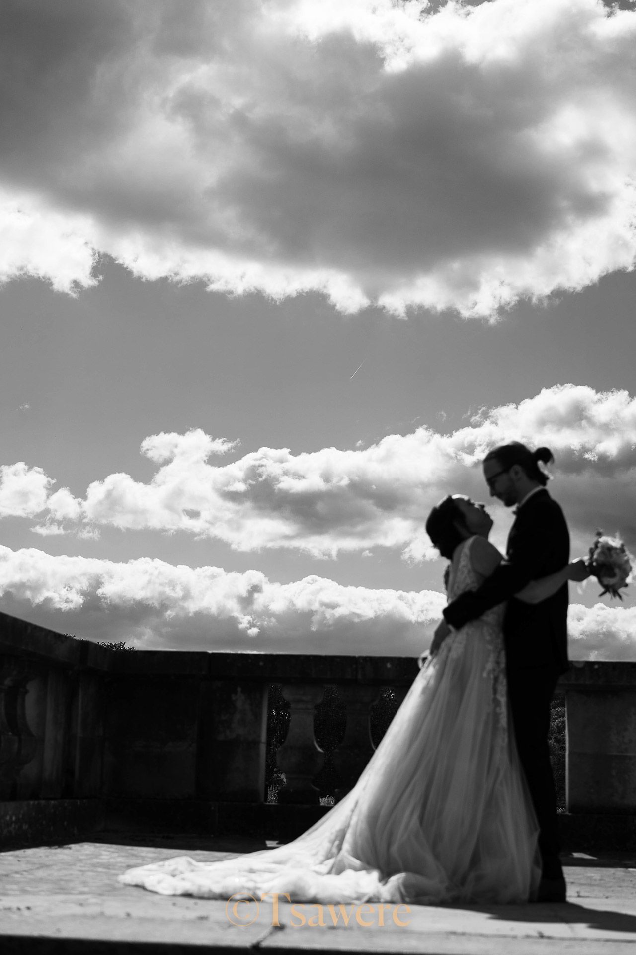 Black and white silhouette of bride and groom embracing under dramatic cloudy sky.