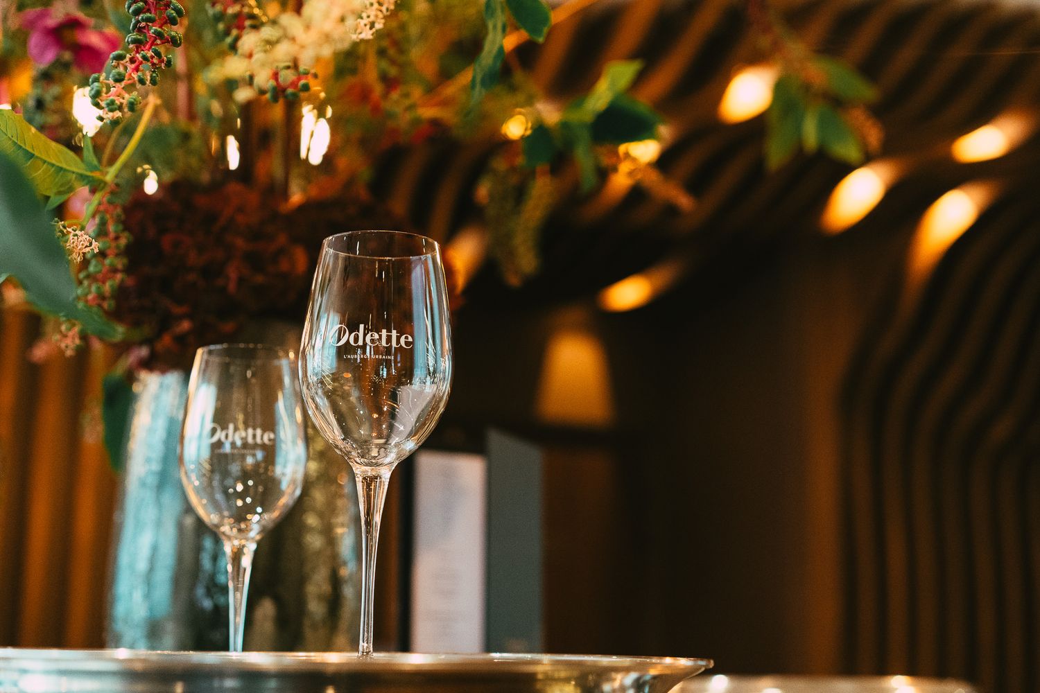 Two Odette branded wine glasses on a table with floral arrangements in a warm restaurant setting.