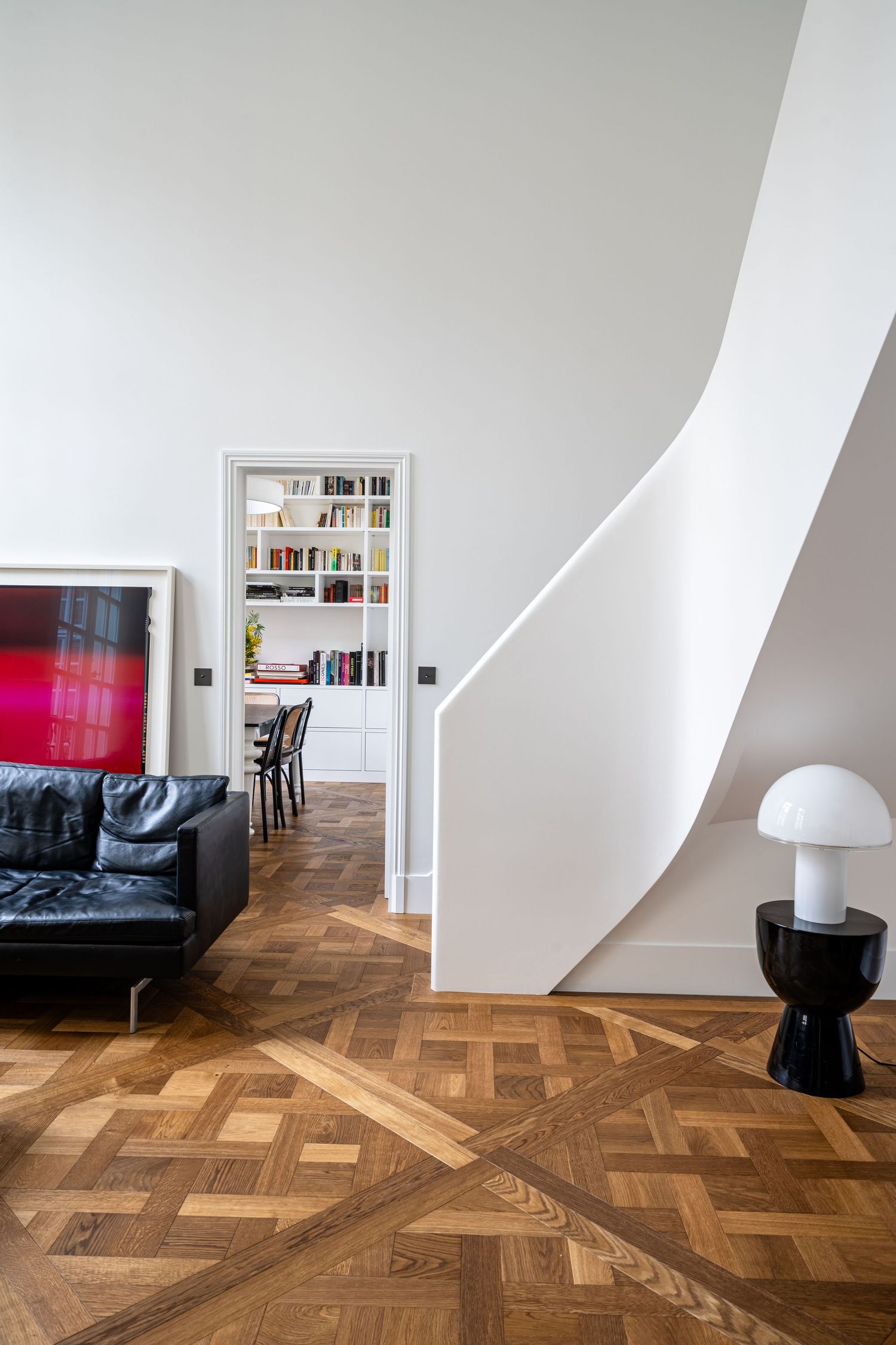 Bright Parisian apartment interior with herringbone parquet floor, curved white staircase, black leather sofa, and red artwork.