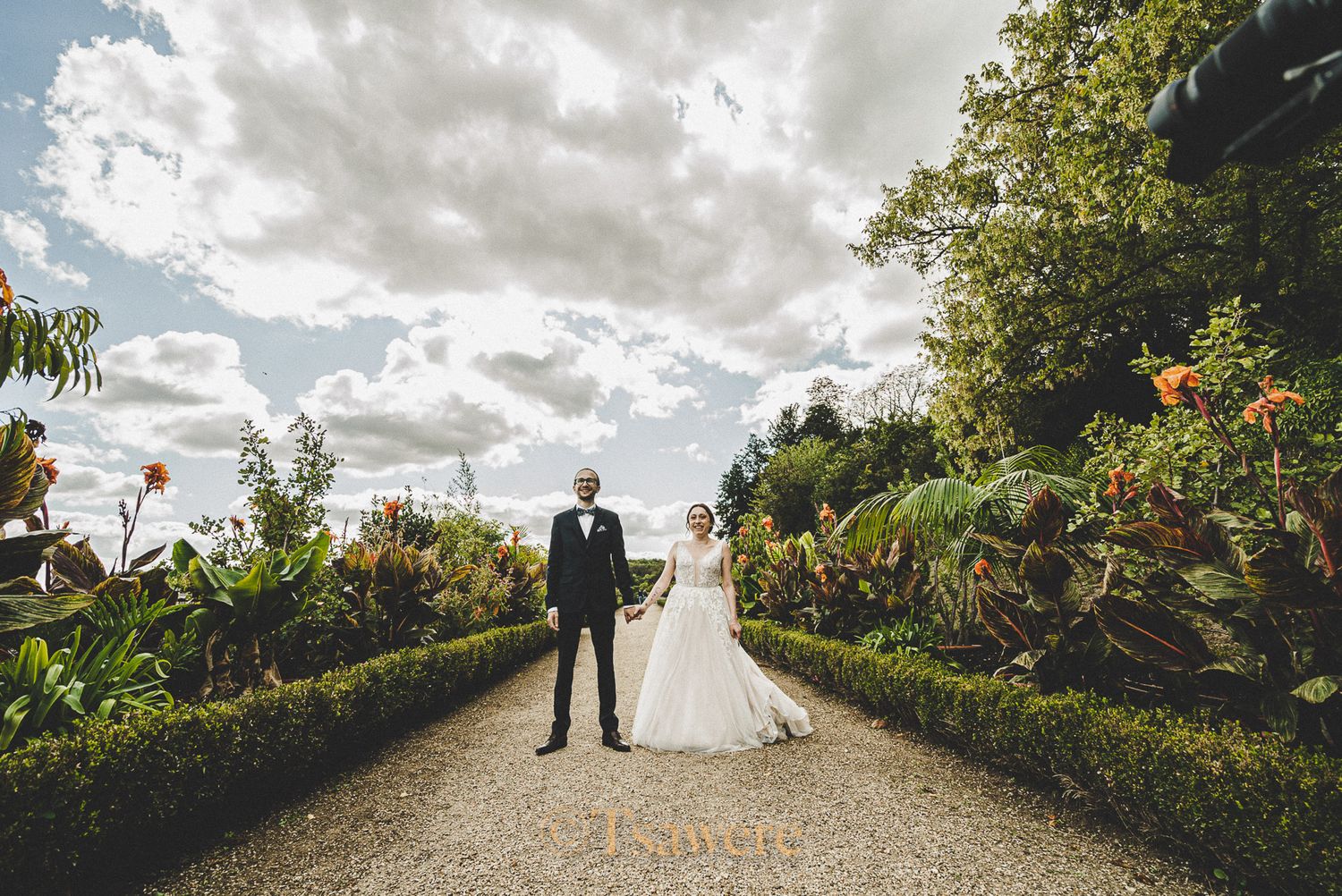 Bride and groom holding hands on garden path surrounded by lush greenery under cloudy sky.