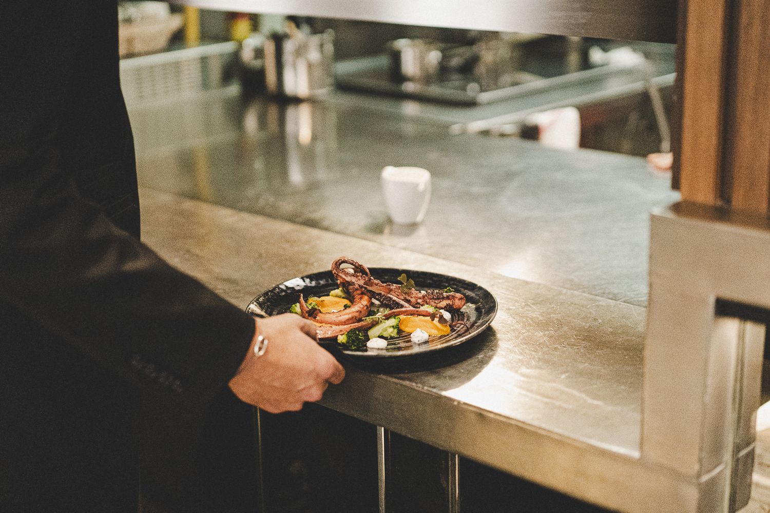 Chef presenting a gourmet octopus dish on a dark plate at a restaurant pass.