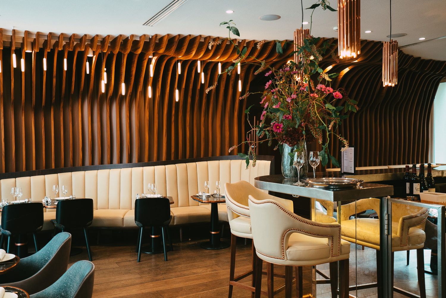 Stylish restaurant dining room with curved wood slat walls, teal velvet booths, pendant lights, and tropical floral display.