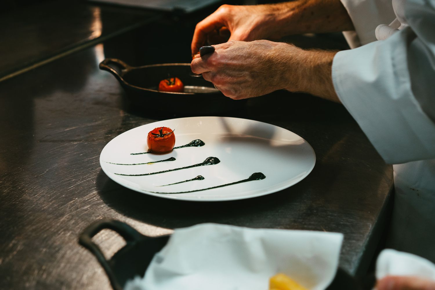 Chef in white coat carefully plates a roasted tomato with decorative herb sauce swirls on a white plate.