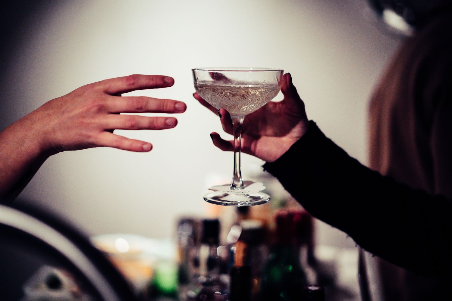 Two hands exchanging a crystal coupe cocktail glass at a bar with bottles in background.