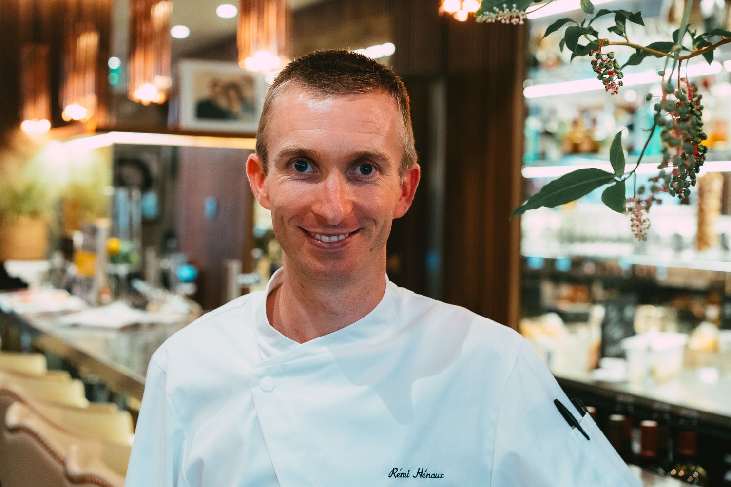 Smiling male chef in white uniform posing in a bright and elegantly decorated restaurant interior.