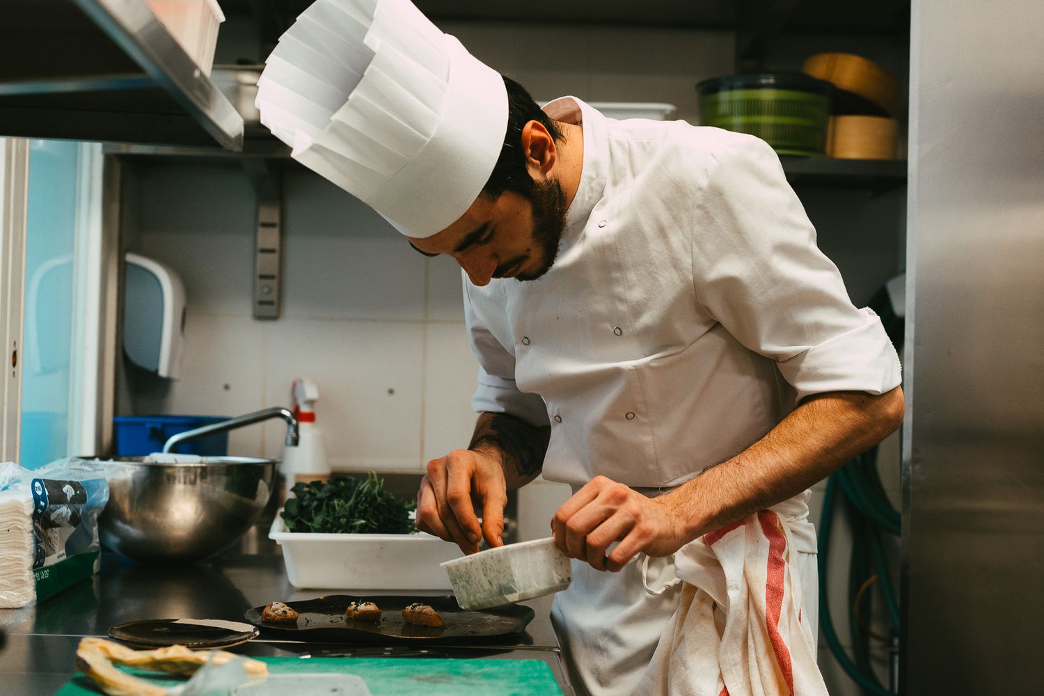 Male chef in white uniform and tall hat carefully garnishing a gourmet dish in a professional kitchen.