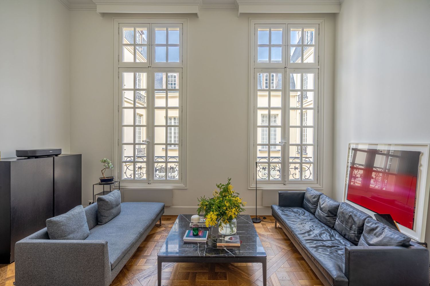 Bright Parisian living room with tall French windows, grey sofas, parquet flooring, and high ceilings.