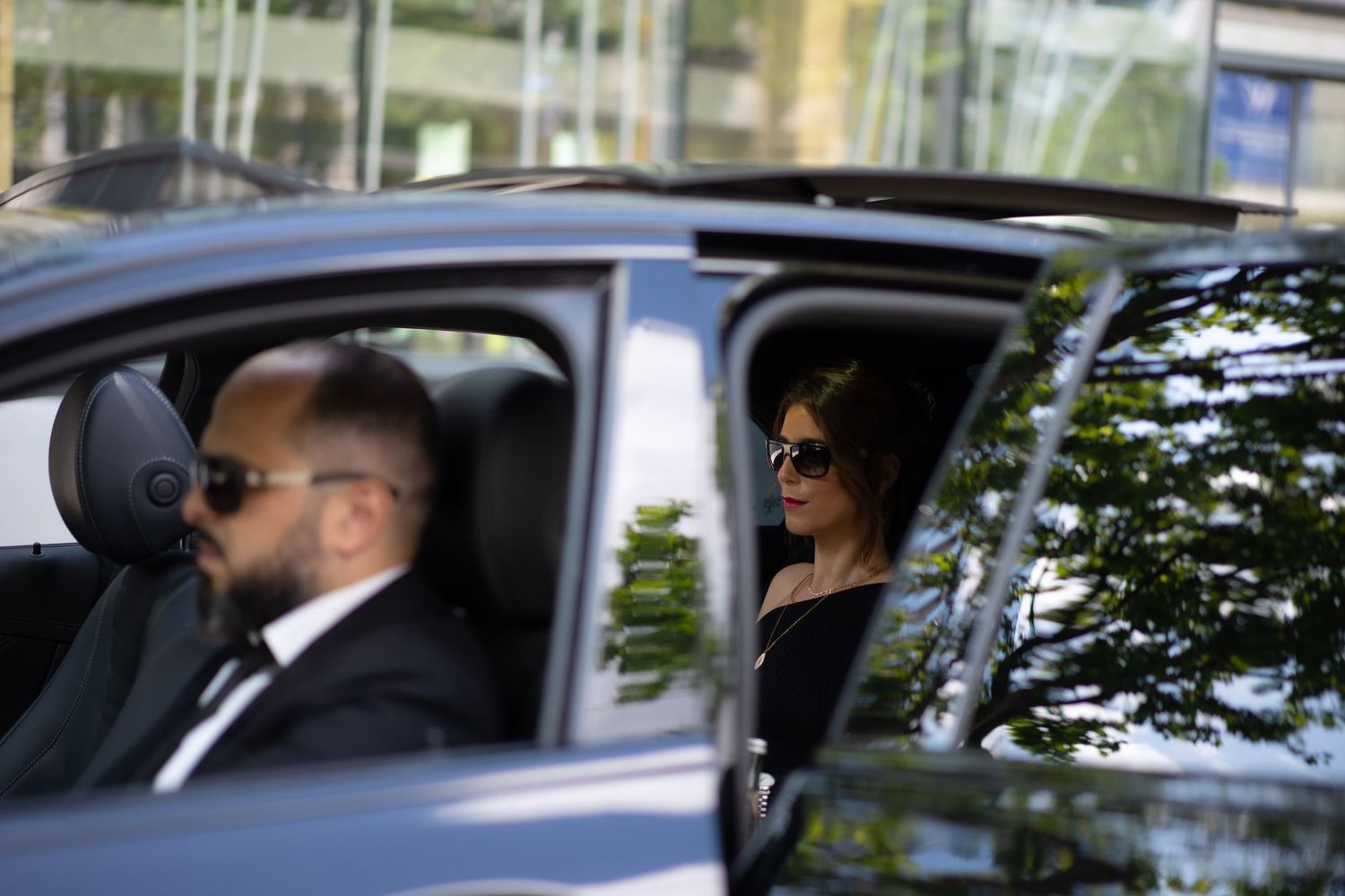 Professional driver and female passenger in sunglasses inside a luxury black vehicle.