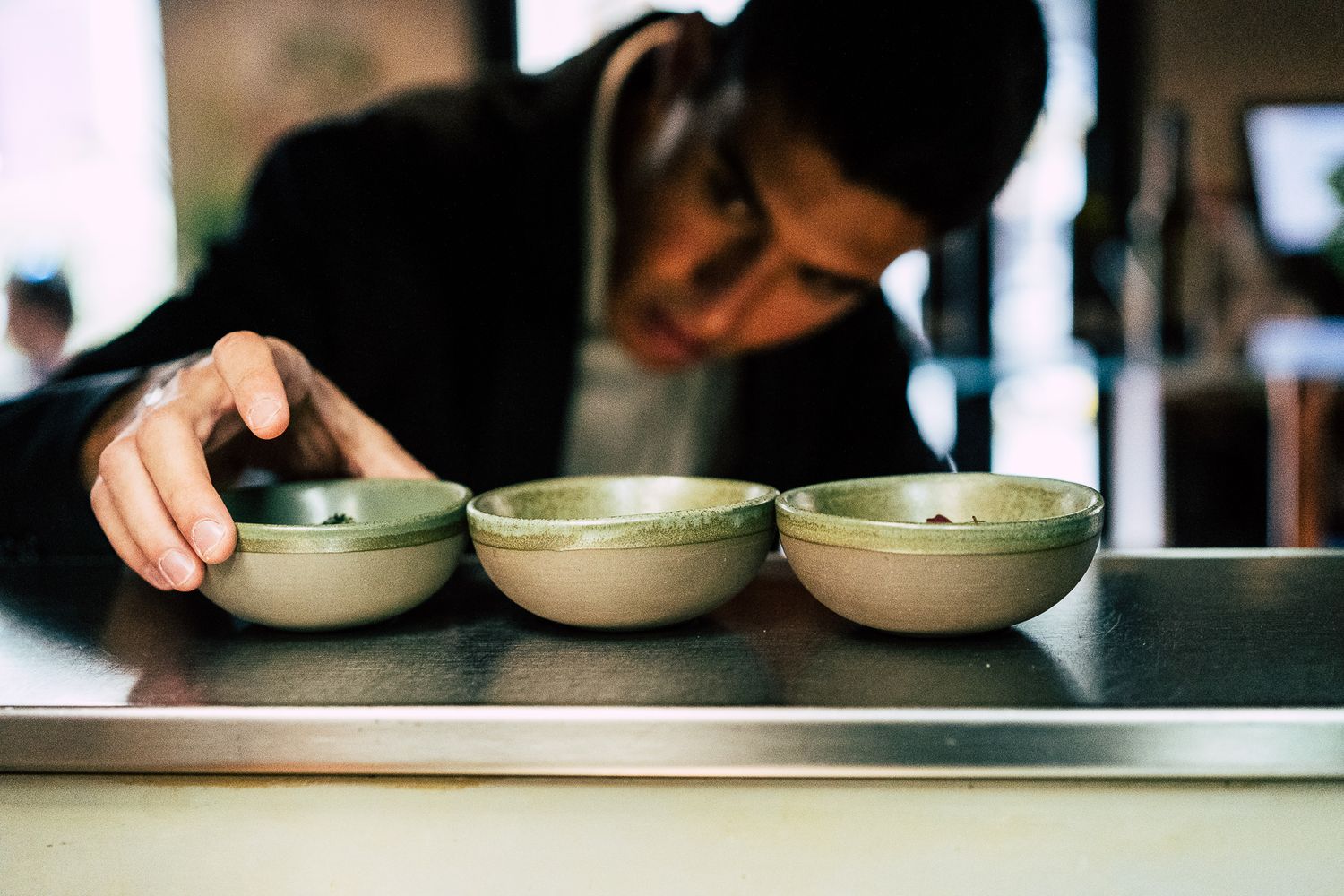 A person arranging three small green ceramic bowls on a stainless steel kitchen counter.