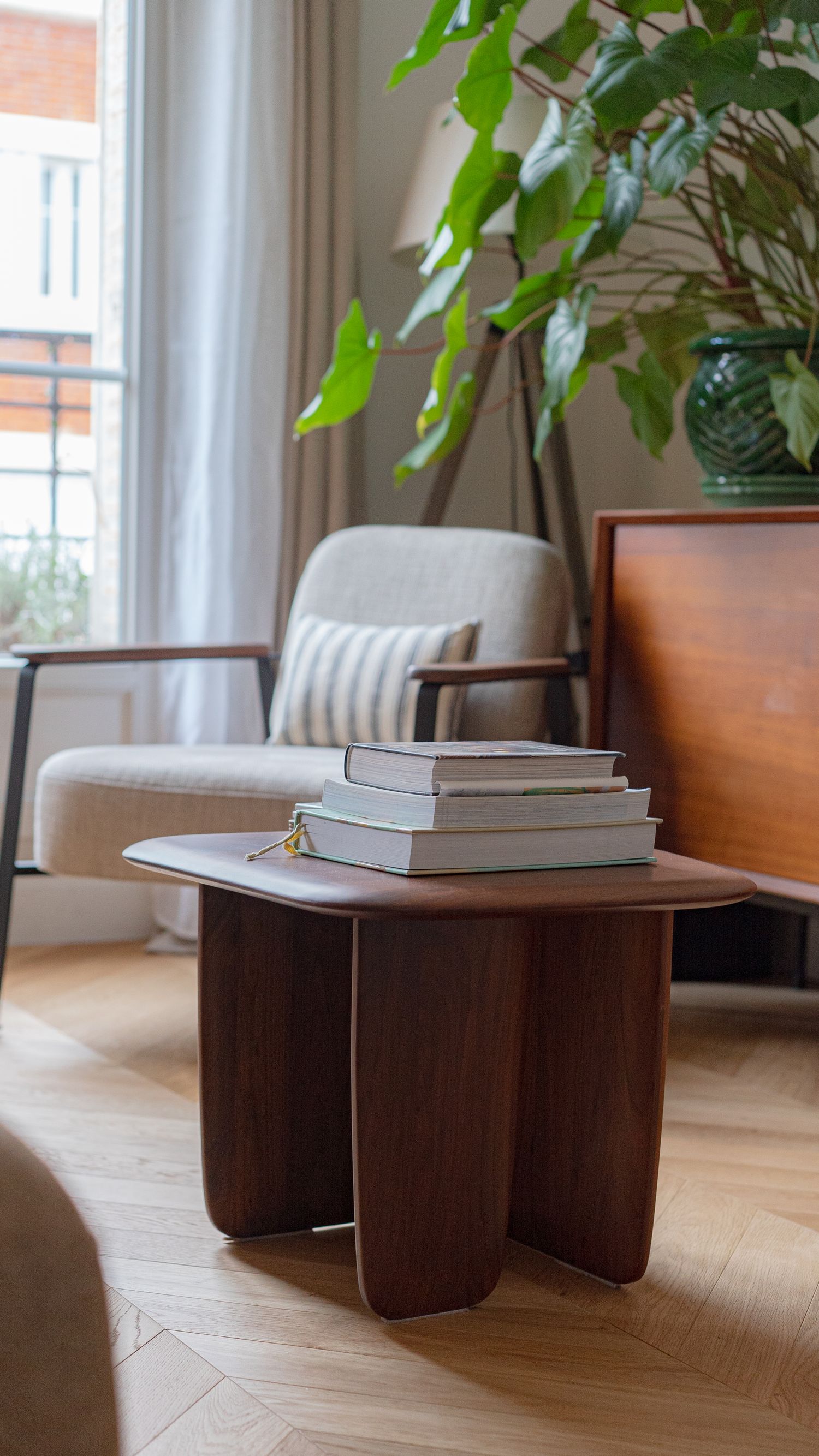 Dark walnut organic-shaped coffee table with stacked books beside a lush green plant and armchair.
