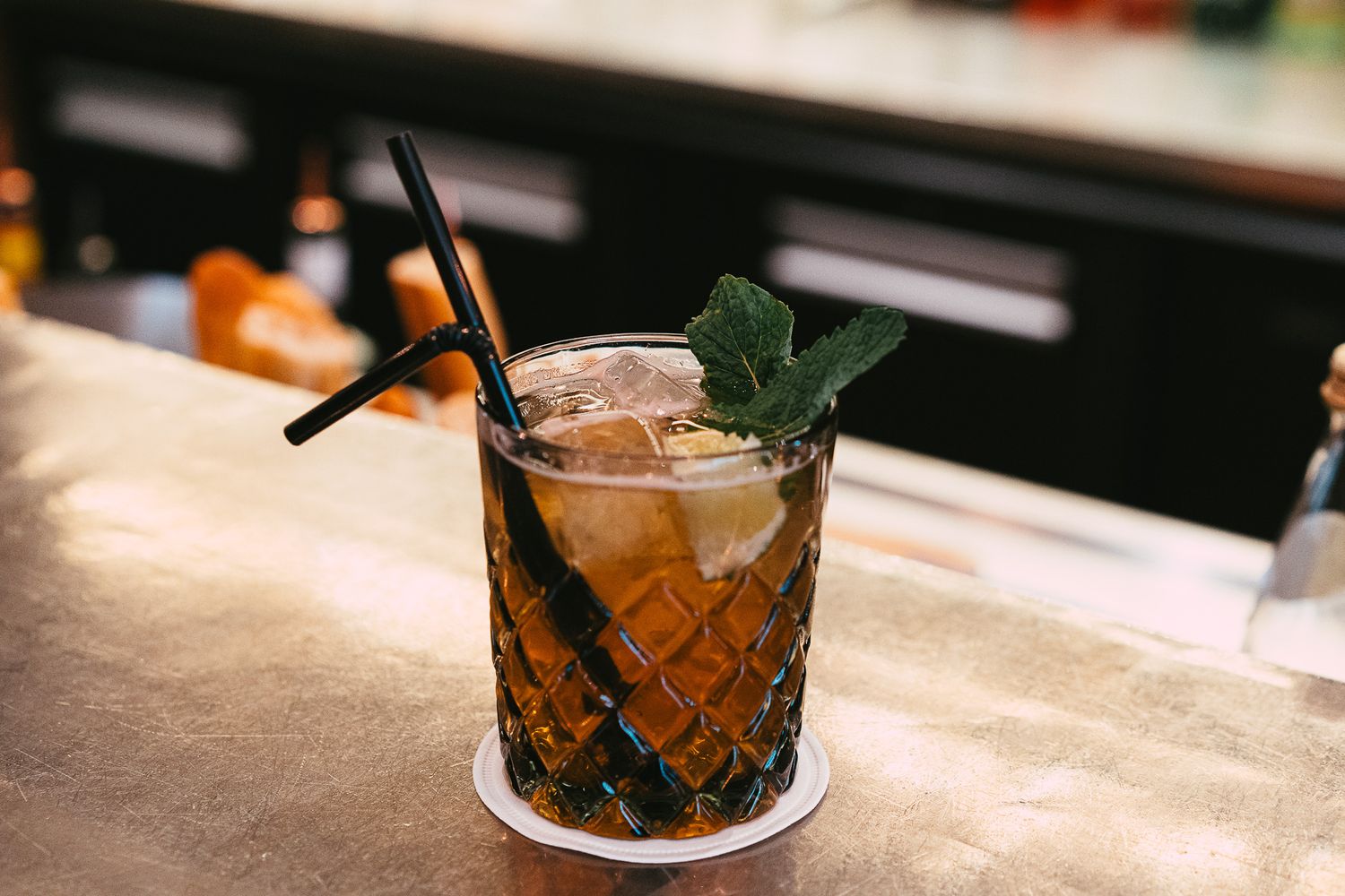 A dark amber cocktail in a crystal glass with ice, mint garnish, and black straws on a bar counter.