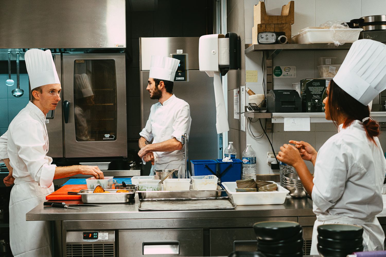 Three chefs in white uniforms and tall hats work together preparing dishes in a busy professional kitchen.
