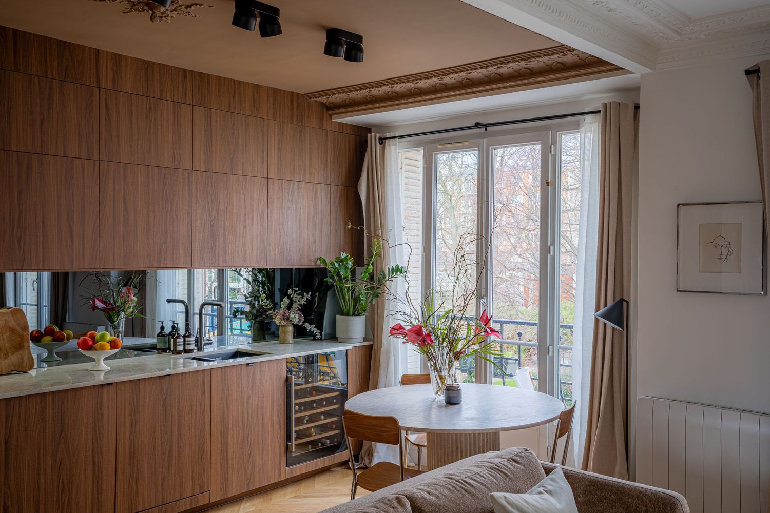 Contemporary walnut kitchen with mirrored backsplash, wine fridge, and round dining table beside large French windows.
