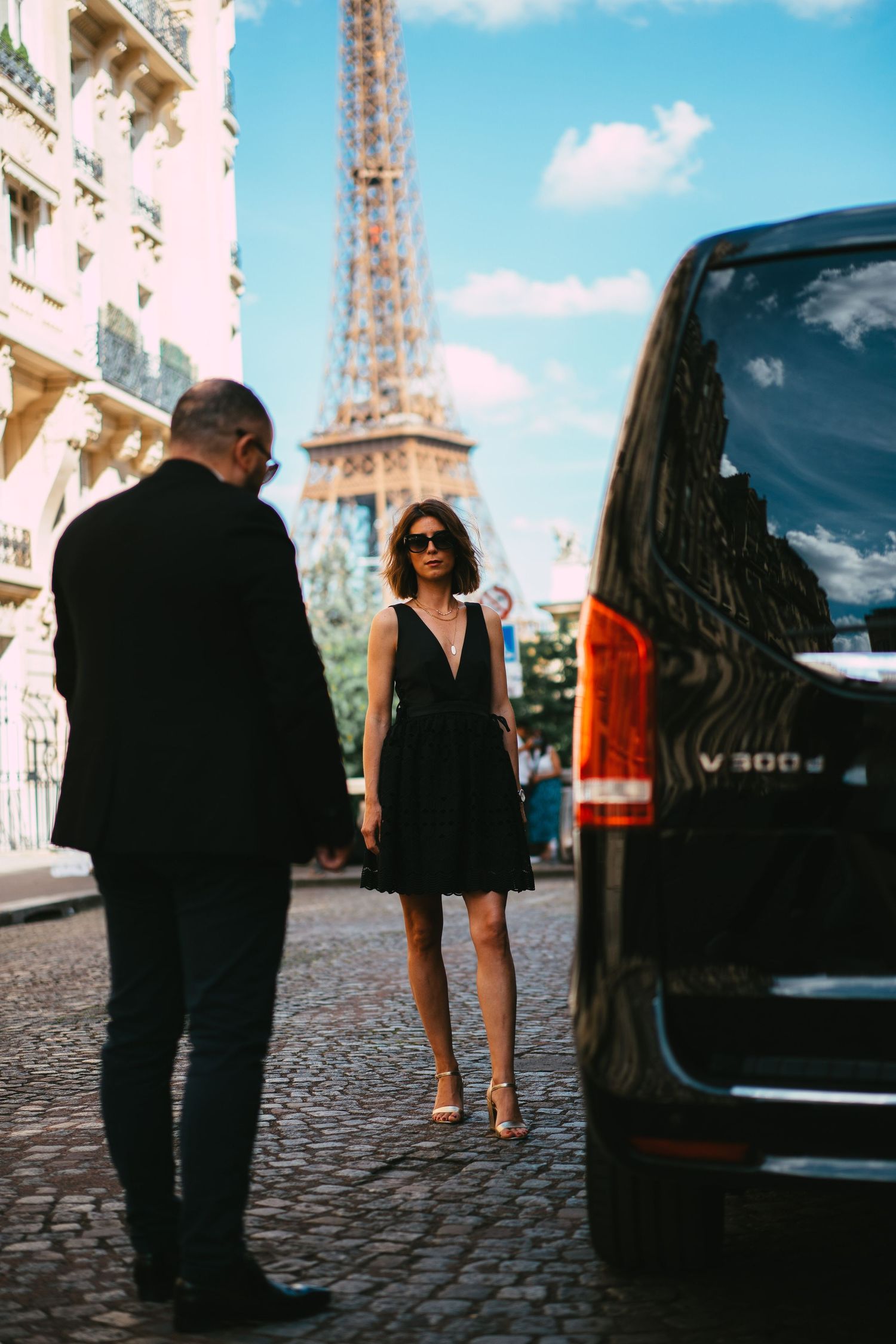 Elegant woman in black dress standing near luxury van with Eiffel Tower backdrop.