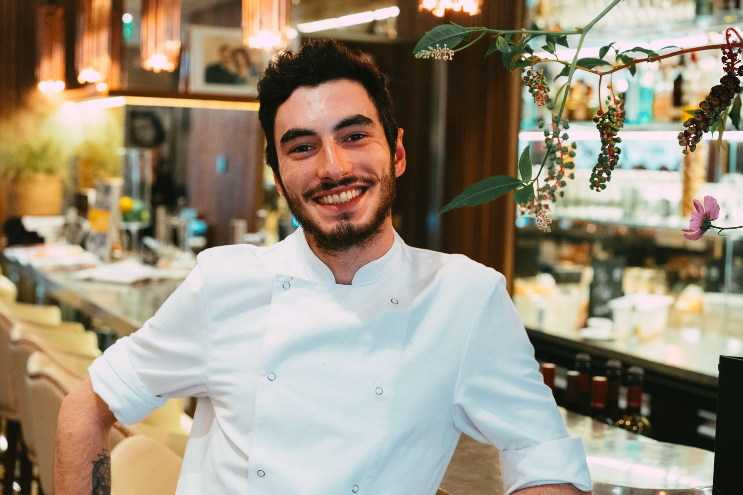 Young smiling male chef in white uniform posing in a beautifully decorated restaurant interior.