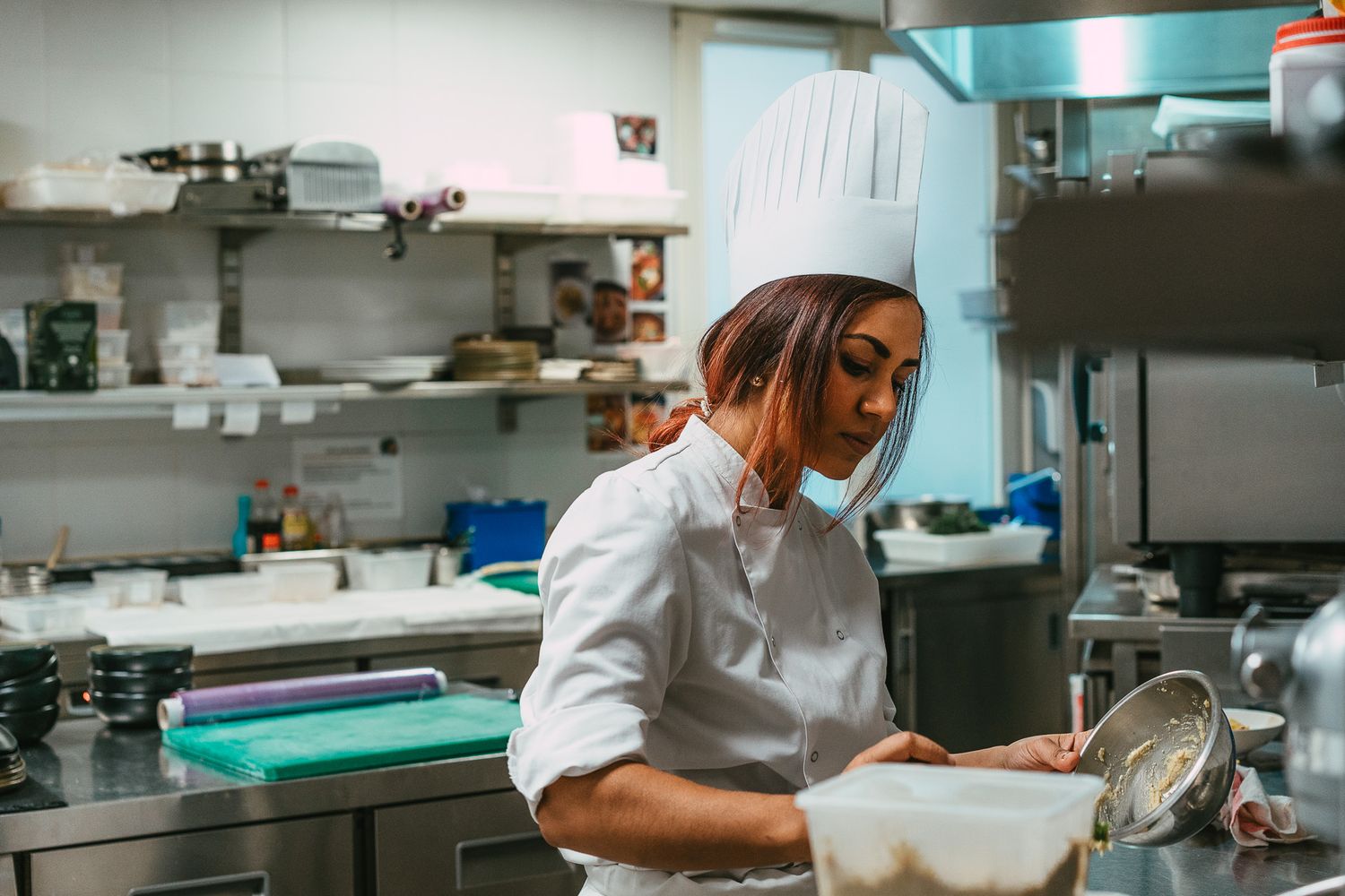 Female chef in white hat and uniform reviewing recipes while holding a bowl in a commercial kitchen.