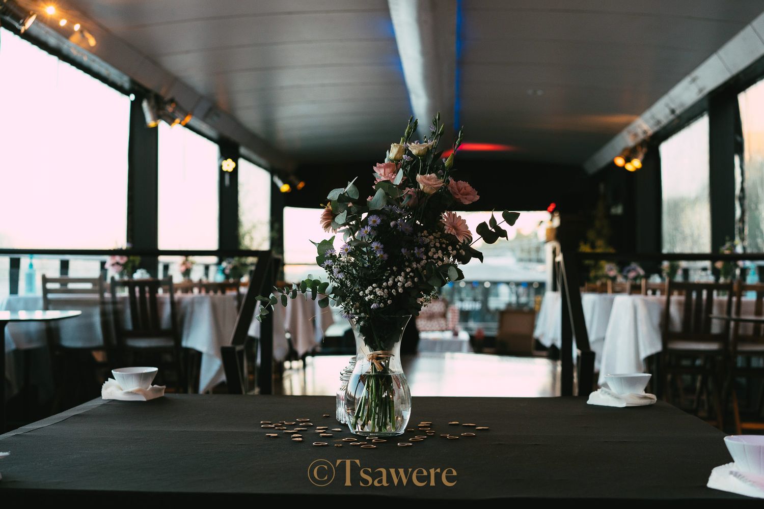 Elegant floral centerpiece on a dark table in a waterfront wedding reception venue at dusk.