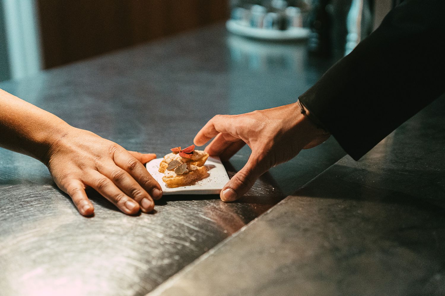 Two hands exchanging a small gourmet appetizer on a white plate over a marble counter.