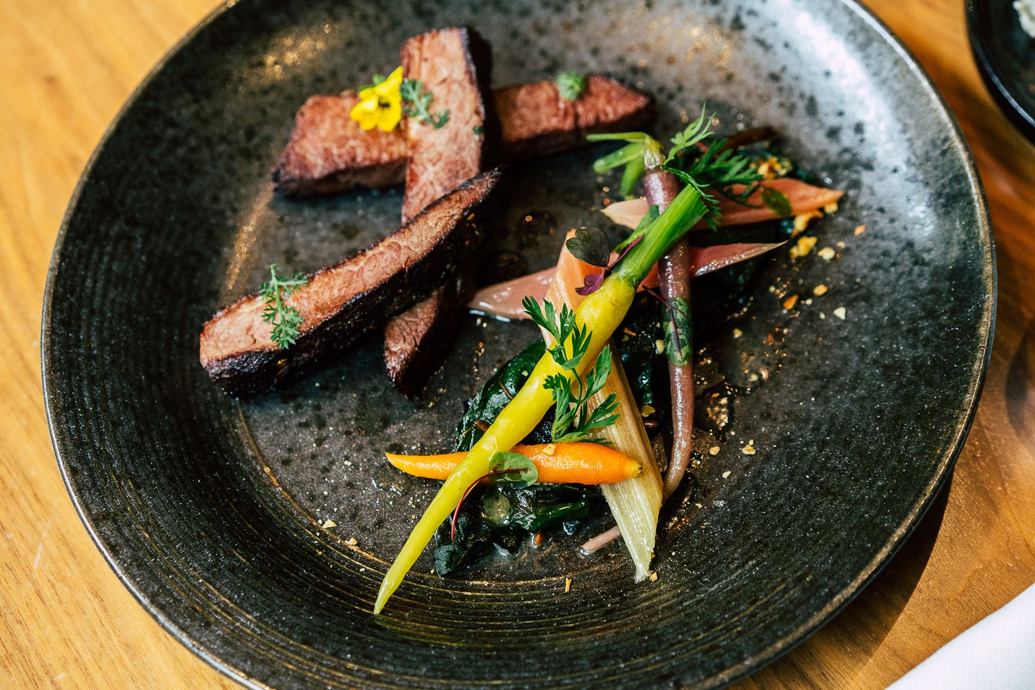Plated sliced beef with colorful roasted vegetables on a dark ceramic plate at a fine dining restaurant.