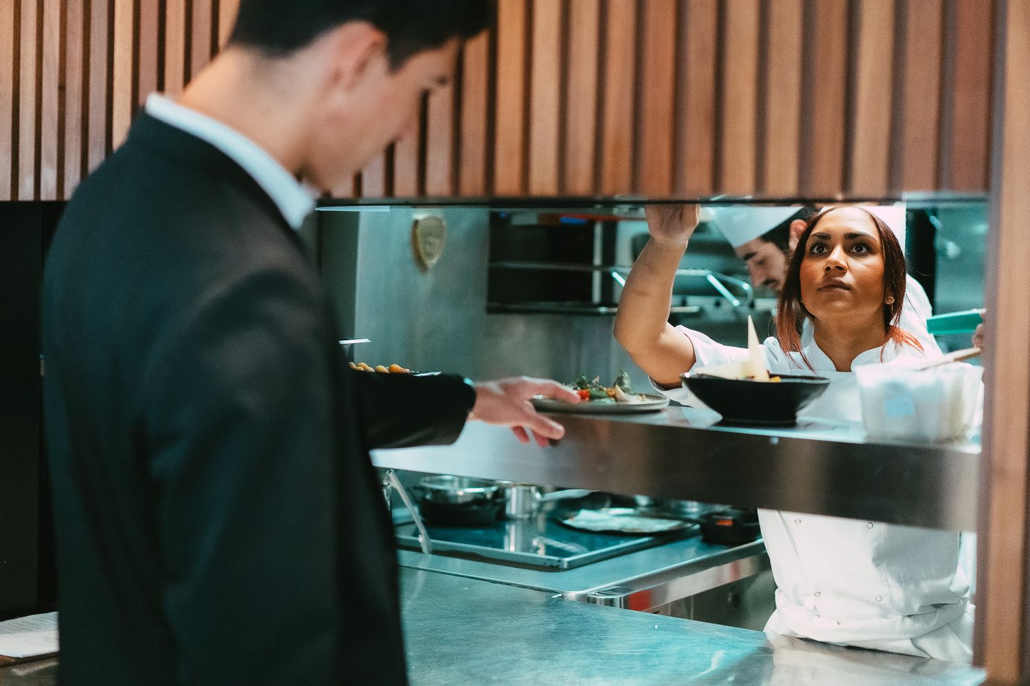 Female chef handing a plated dish to a waiter through a restaurant kitchen pass window.