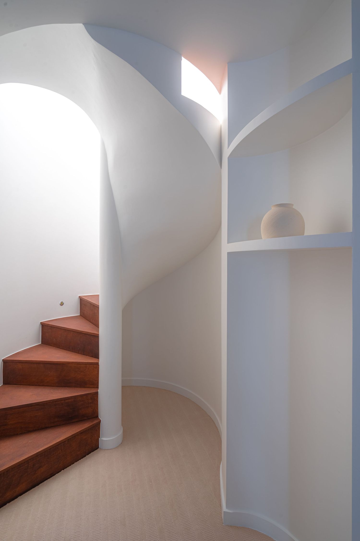 Curved white spiral staircase with warm wooden steps, arched alcove, and built-in shelving niche.
