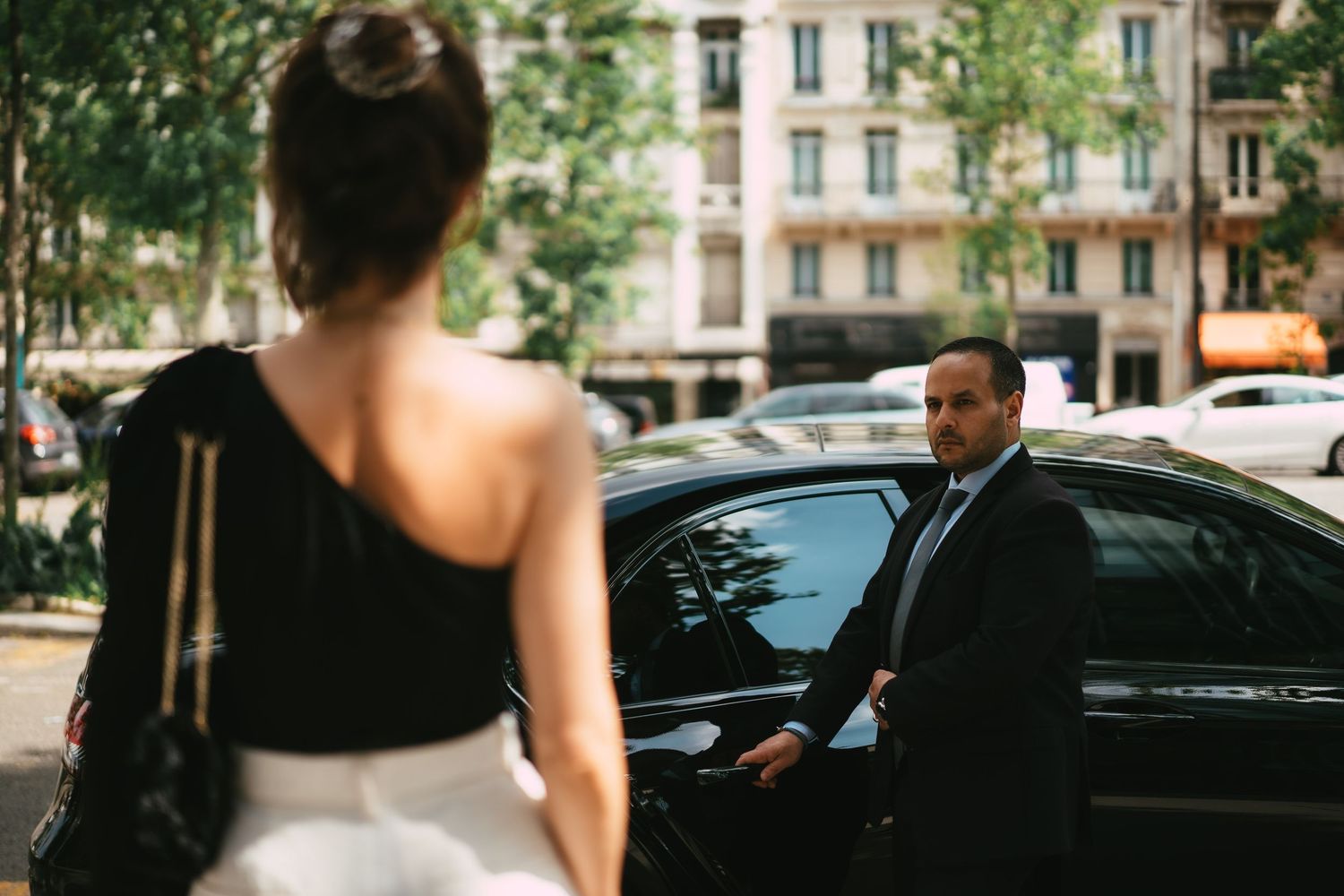 Professional chauffeur opening car door for elegantly dressed woman on city street.