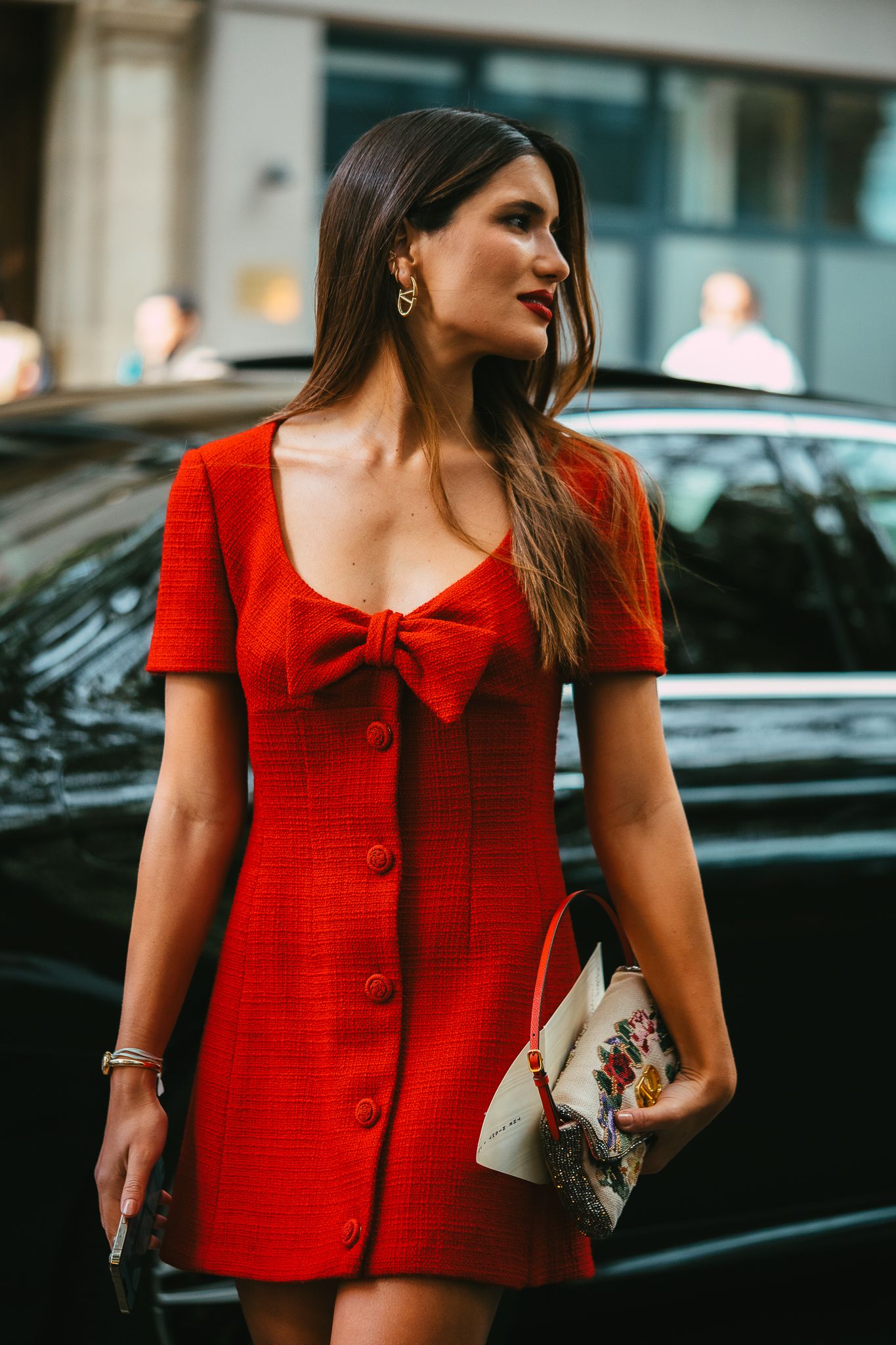 Brunette woman in red bow mini dress holding floral clutch at fashion week street style.