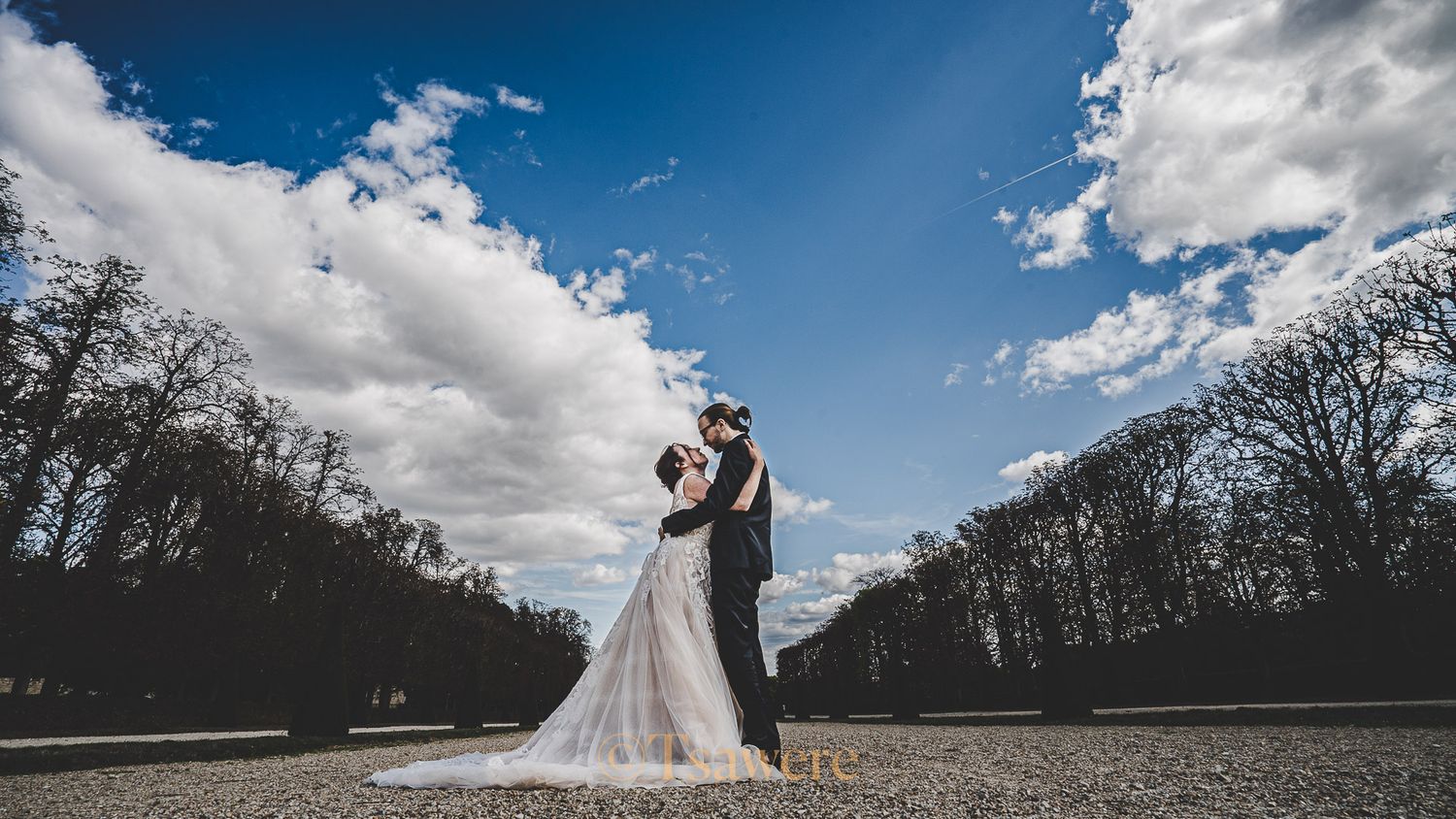 Bride and groom embracing on gravel path lined with trees under a beautiful blue sky.