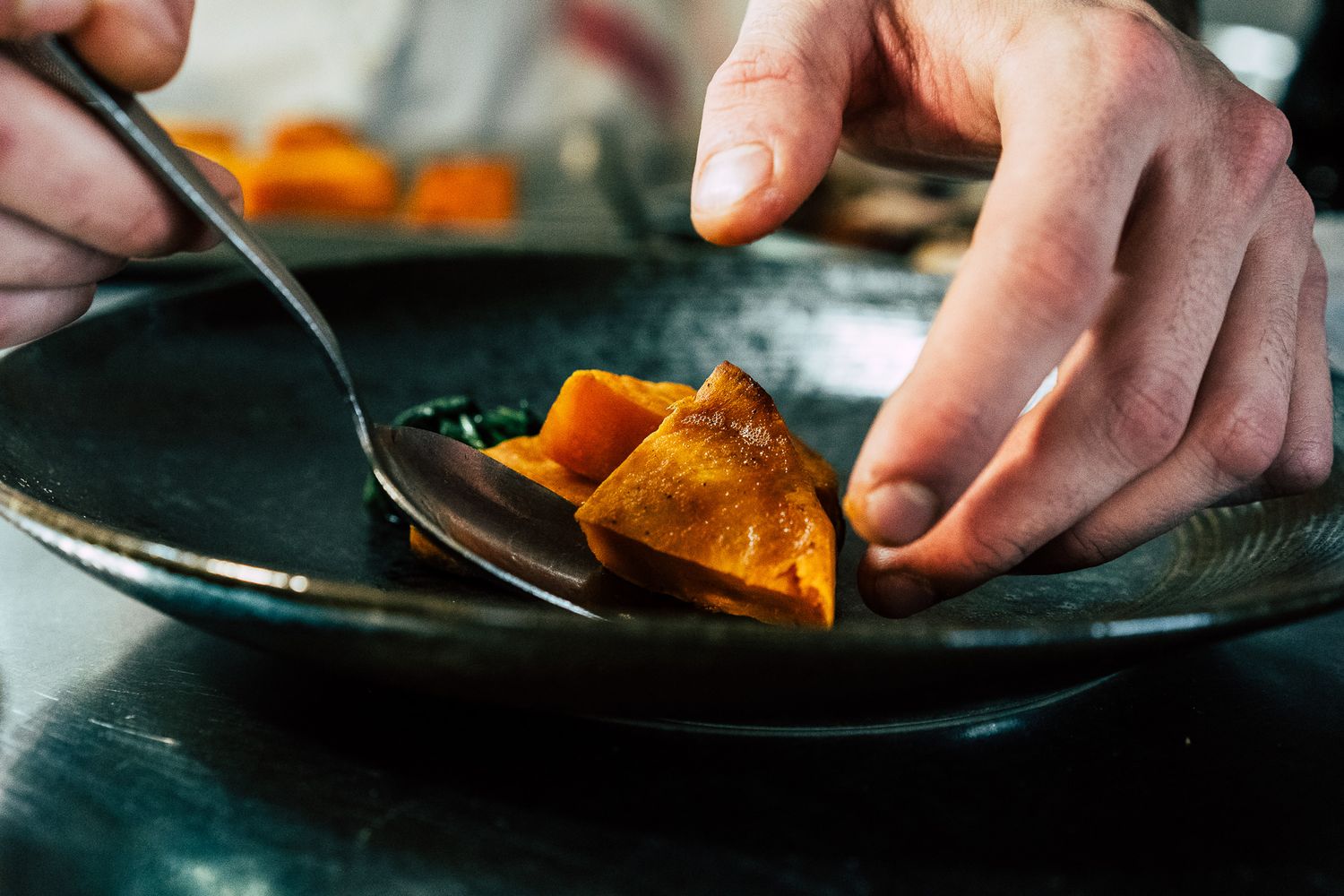 Chef plating roasted pumpkin on a dark ceramic dish with a spoon in a professional kitchen.