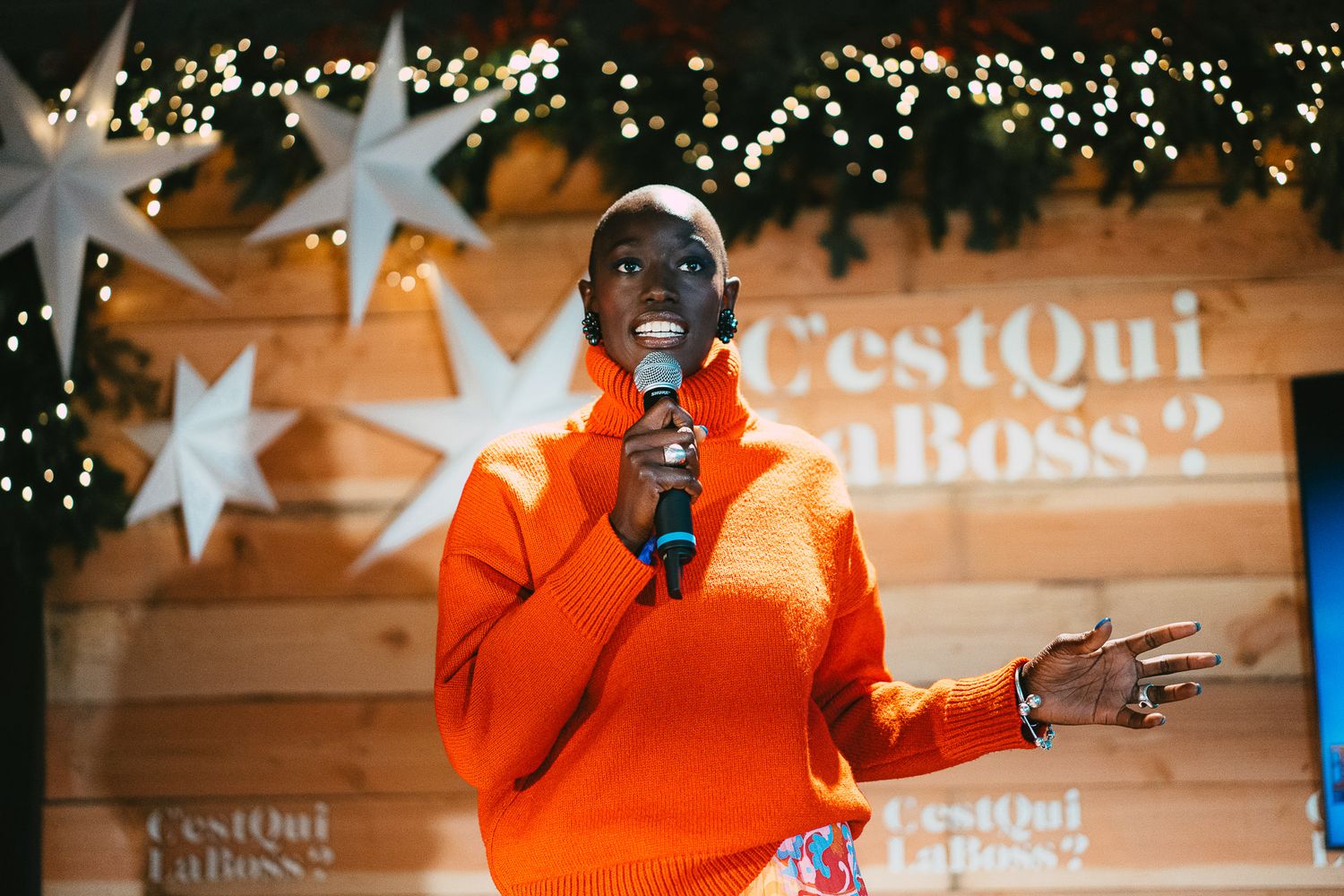 Woman in orange turtleneck sweater speaking into microphone at C'est Qui La Boss event.